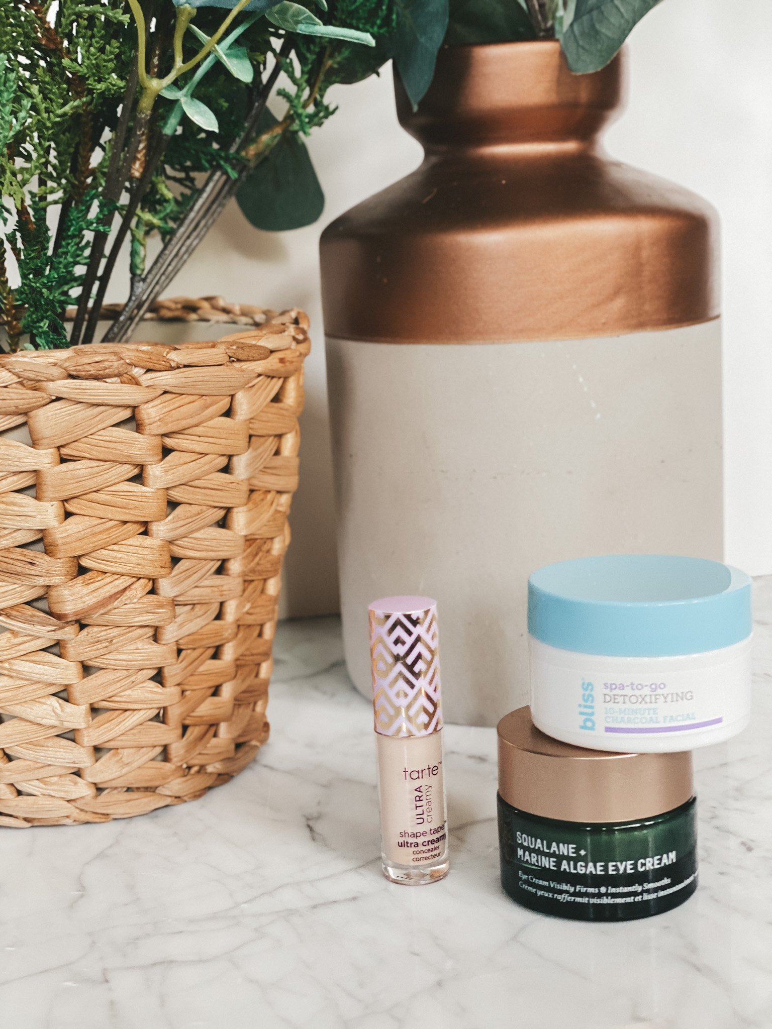 a marble table with 2 small jars with an eye cream and face mask and a small tube on concealer next to them; a cement and bronze vase and a woven basket with a faux potted plant