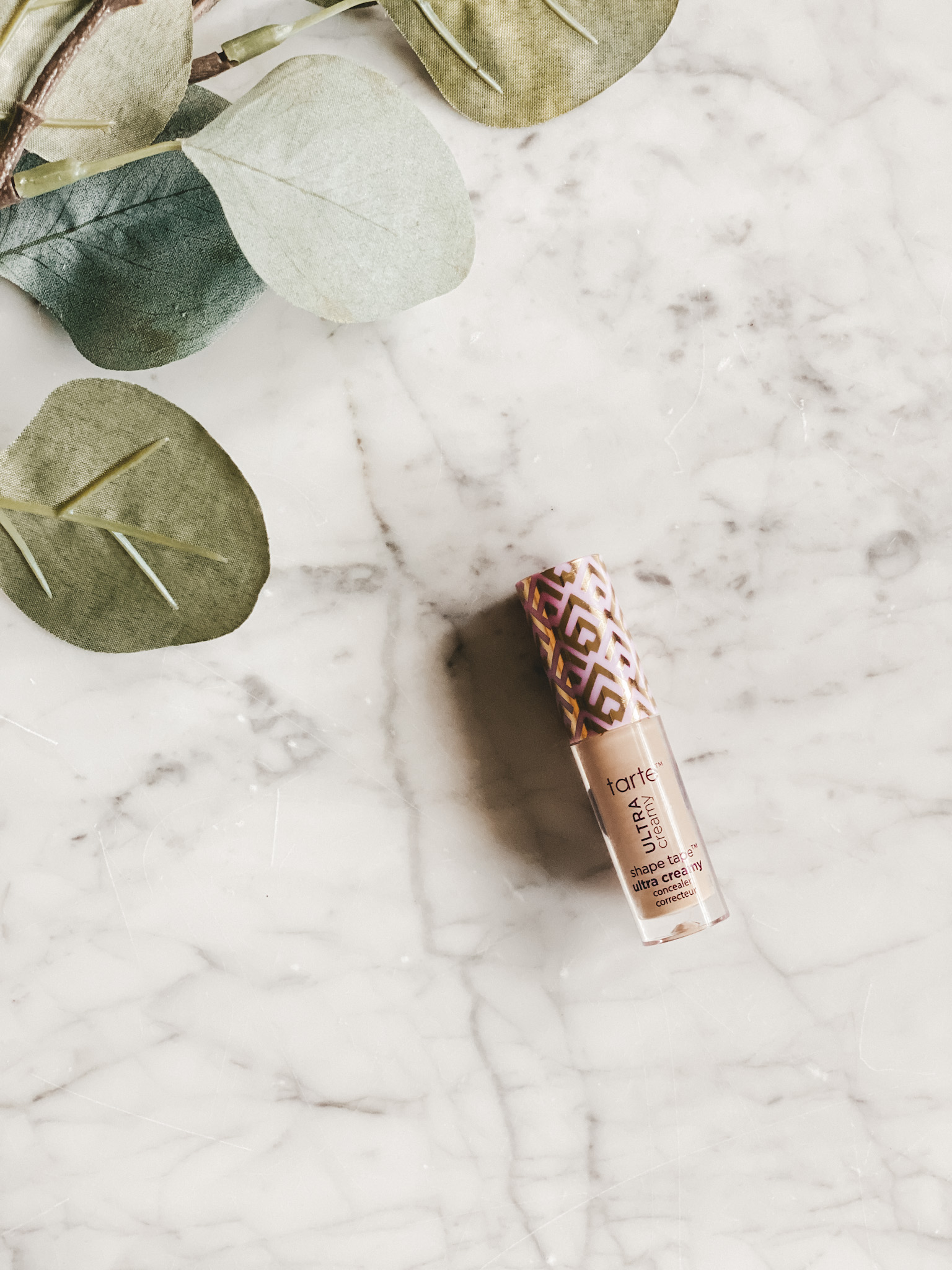 a bottle of Tarte Shape Tape Creamy Concealer set on a marble table with a eucalyptus leaves in the corner