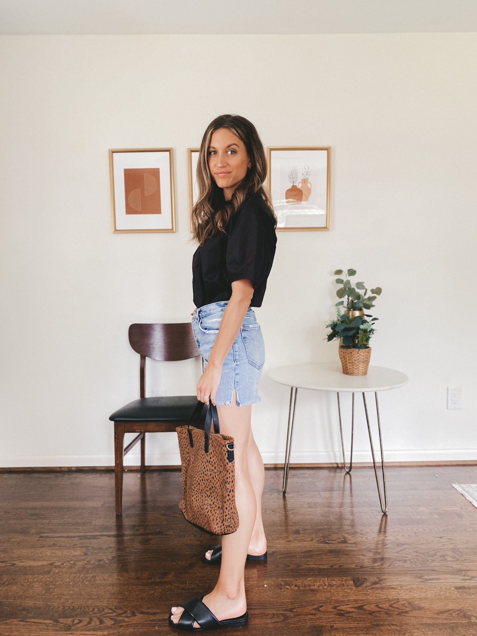 ashley wearing a black short sleeved top, denim cut-off shorts, black crossover slides and a leopard tote bag