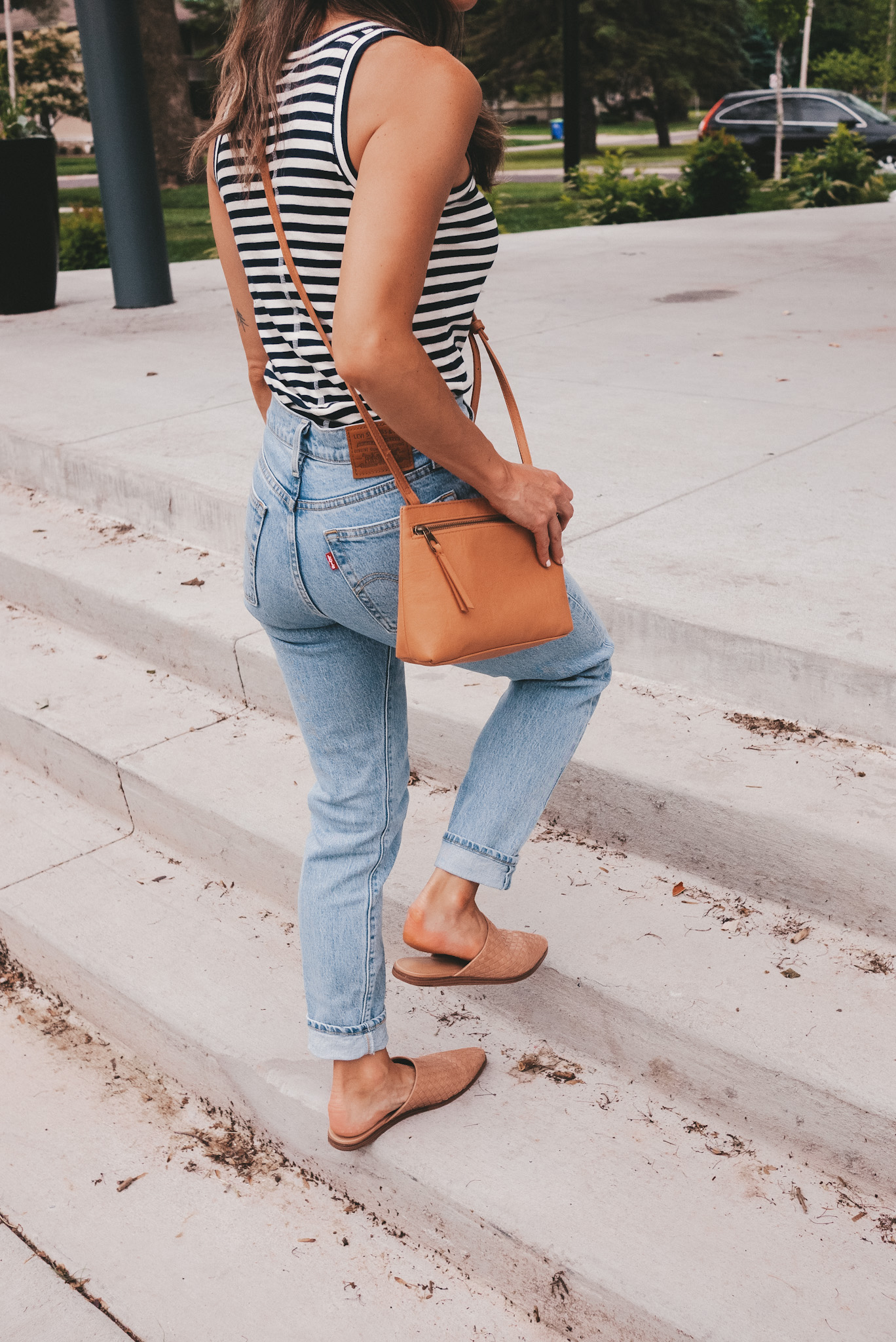 ashley wearing a navy and white striped tank, light wash mom jeans and tan woven mules with a camel colored crossbody purse walking up a set of concrete stairs