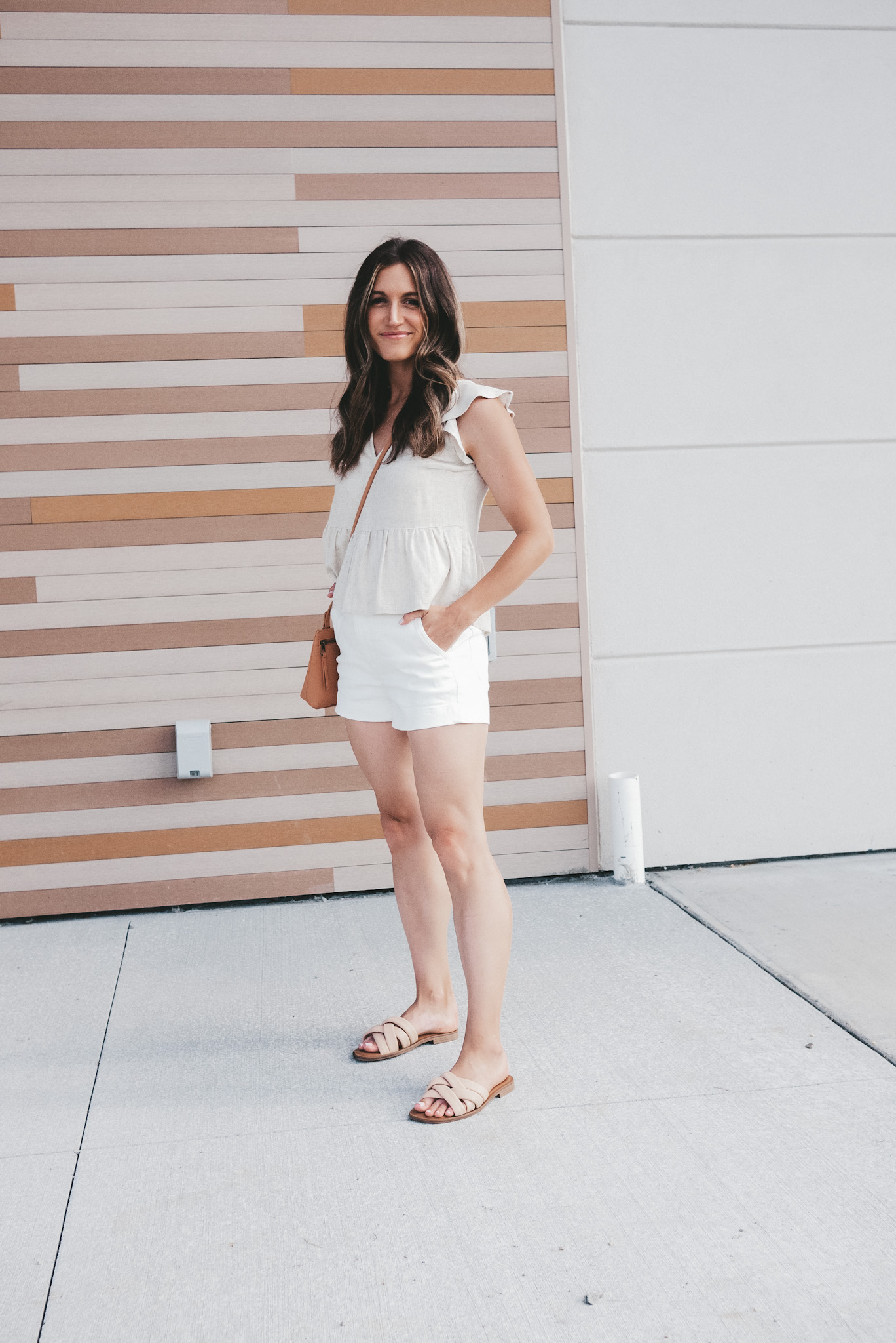 ashley wearing a tan flutter sleeve top, white twill shorts, nude padded sandals and a camel colored crossbody bag standing next to a building
