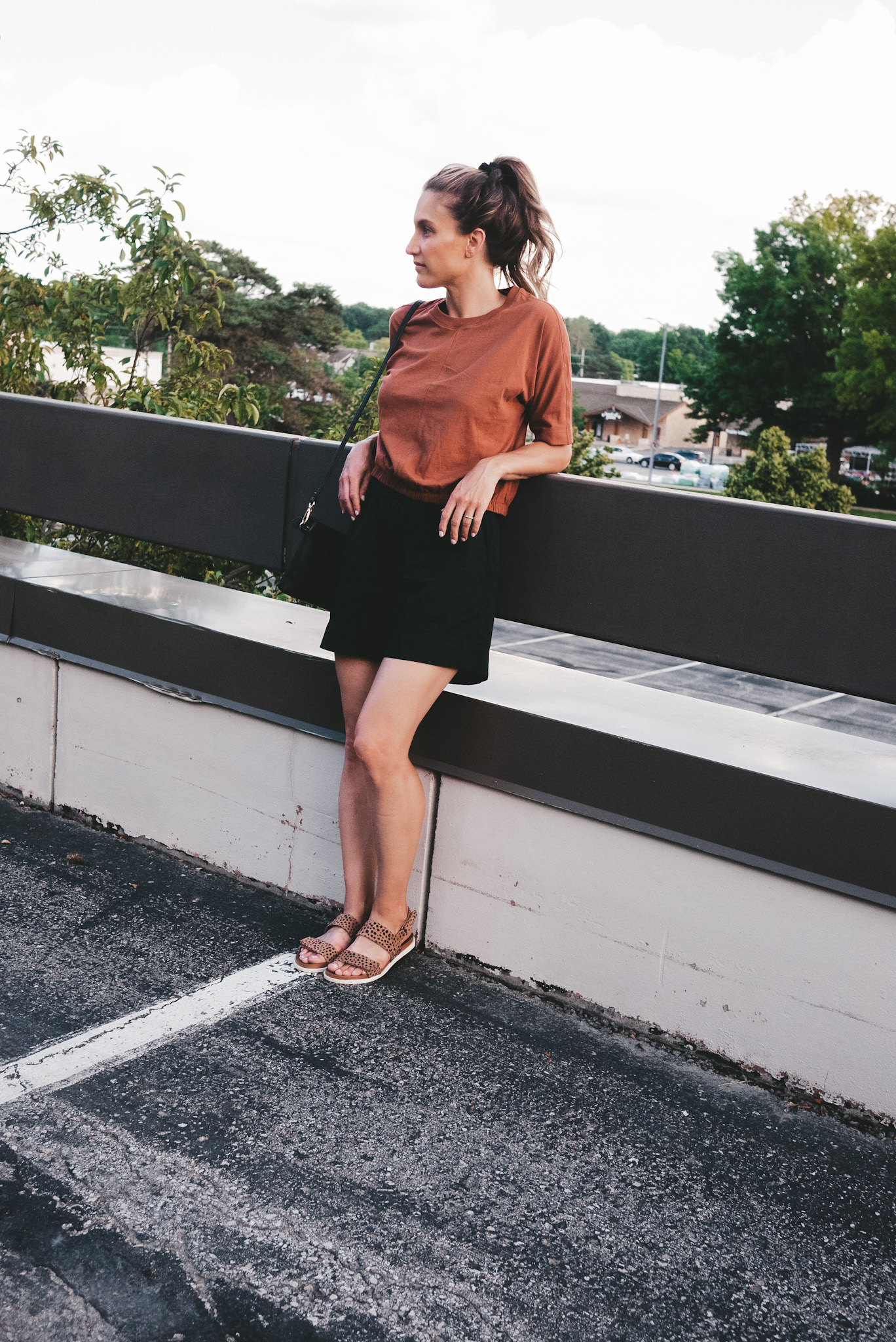 ashley wearing a rust colored tee, black chino shorts and leopard print sandals leaning backward on a rail in a parking lot