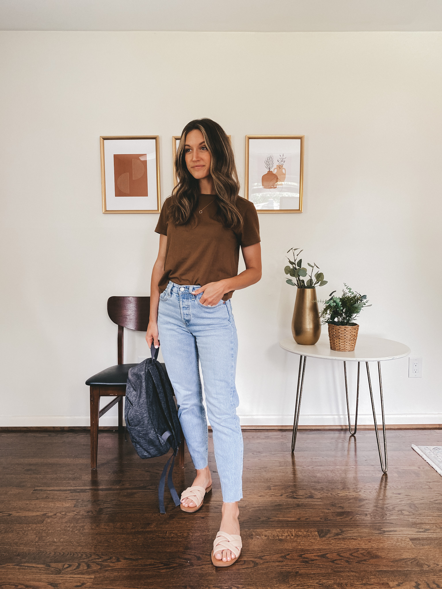 ashley wearing a walnut colored tee, light wash straight leg denim and nude padded slides holding a dark gray backpack