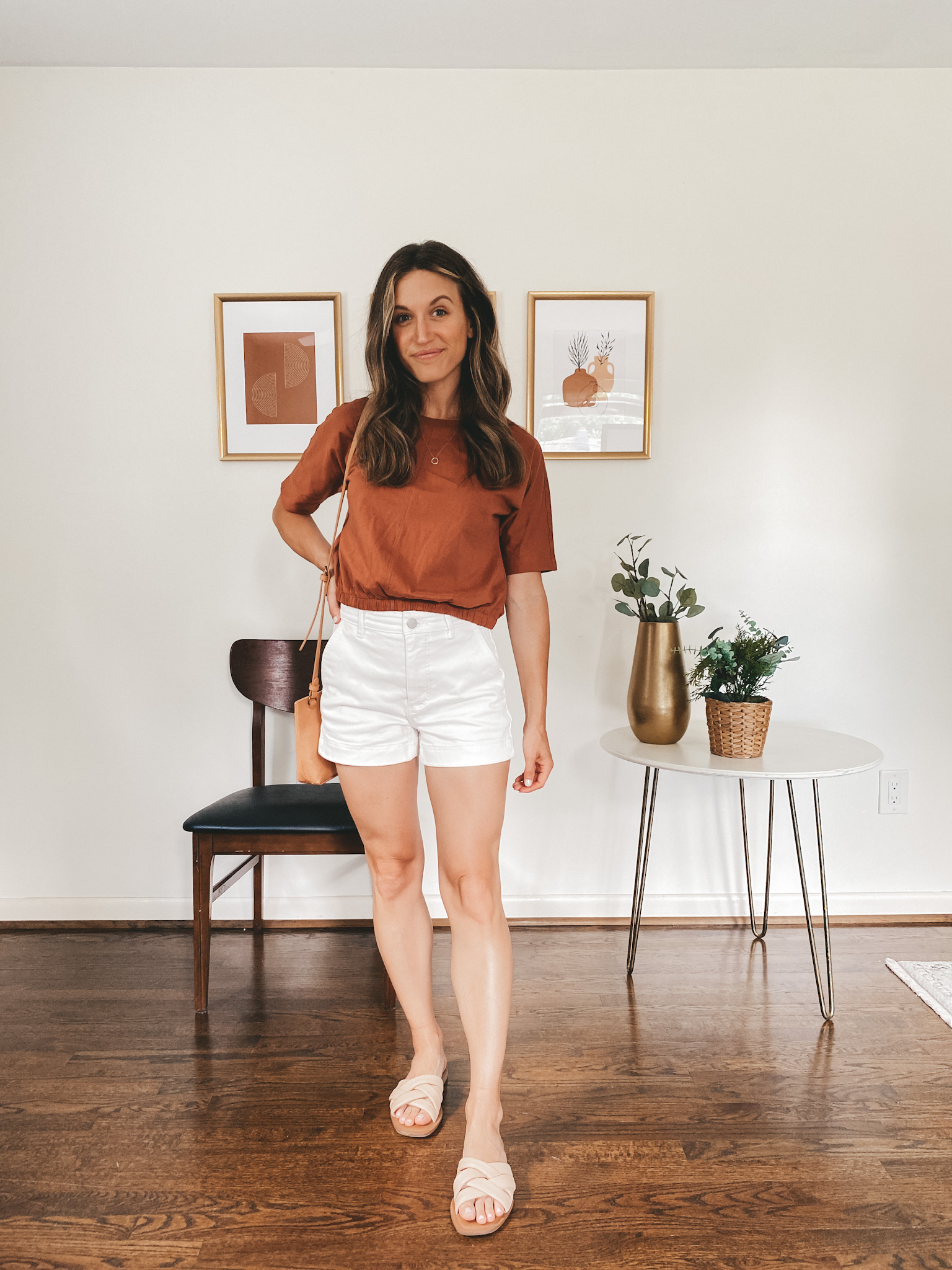 ashley wearing a rust colored bubble tee, white chino shorts and nude padded slides with a camel colored crossbody bag