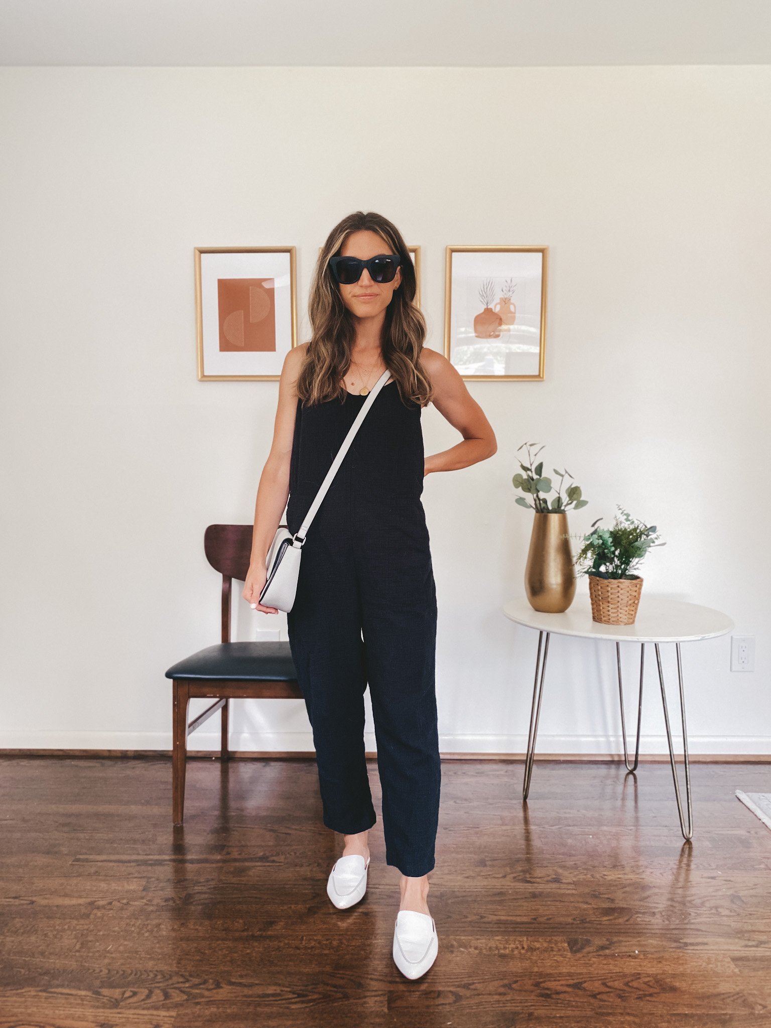 ashley wearing a black jumpsuit, white mules, black square frame sunglasses and a gray crossbody bag