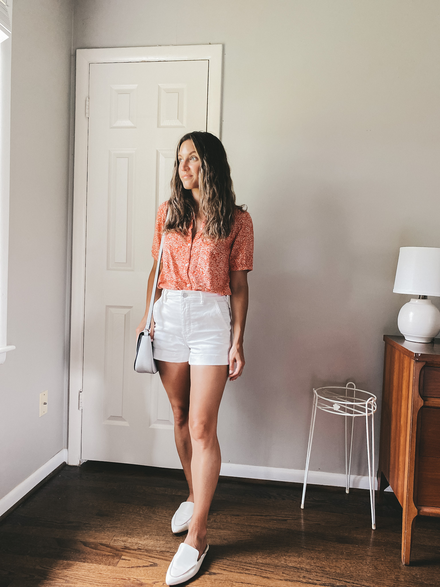 ashley wearing a red button down floral blouse, white twill shorts and white mules with a gray crossbody bag
