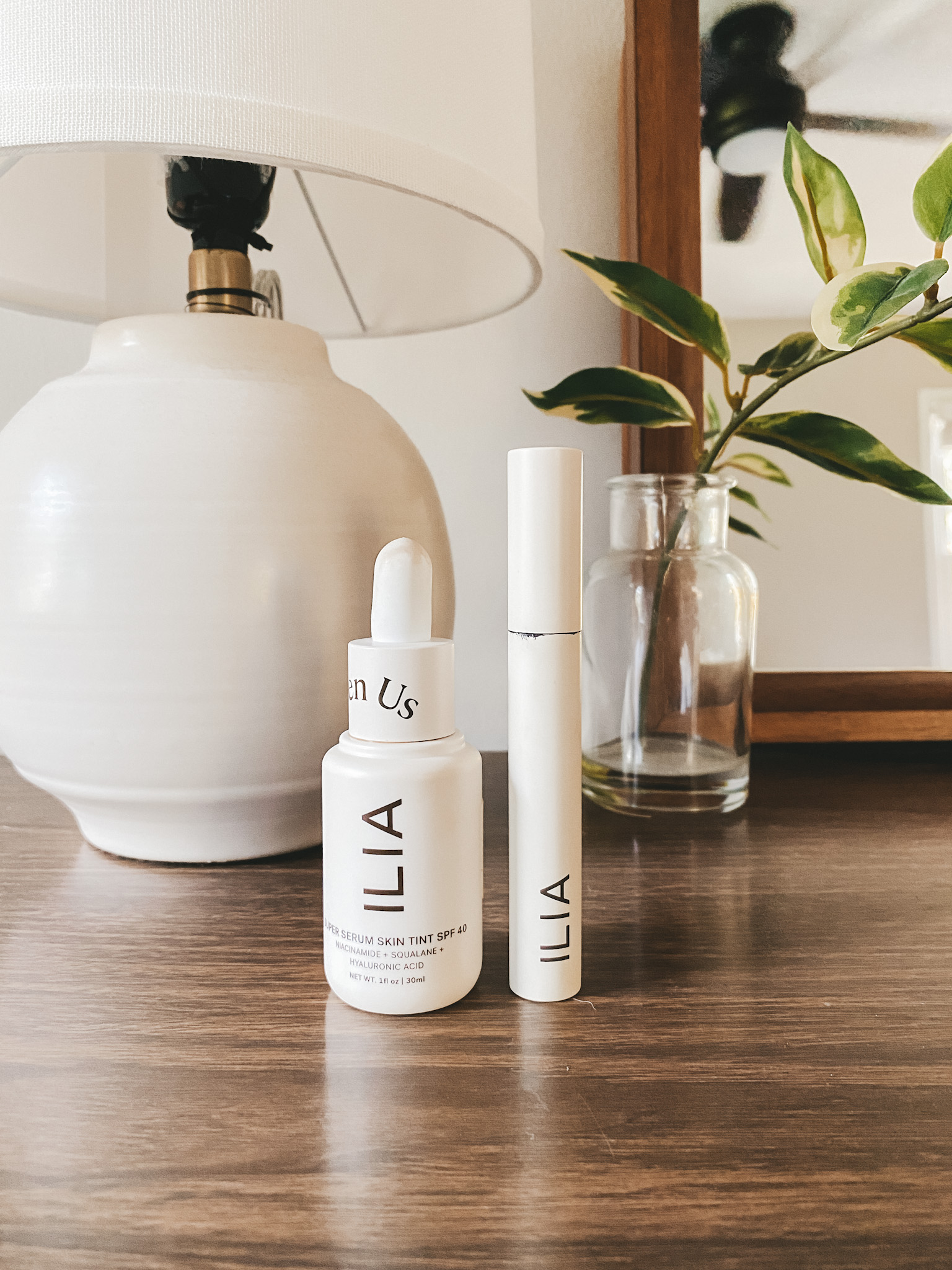 a bottle of Ilia Serum Foundation and Ilia Mascara on a vintage wooden dresser with a white table lamp and single plant stem in a clear glass vase
