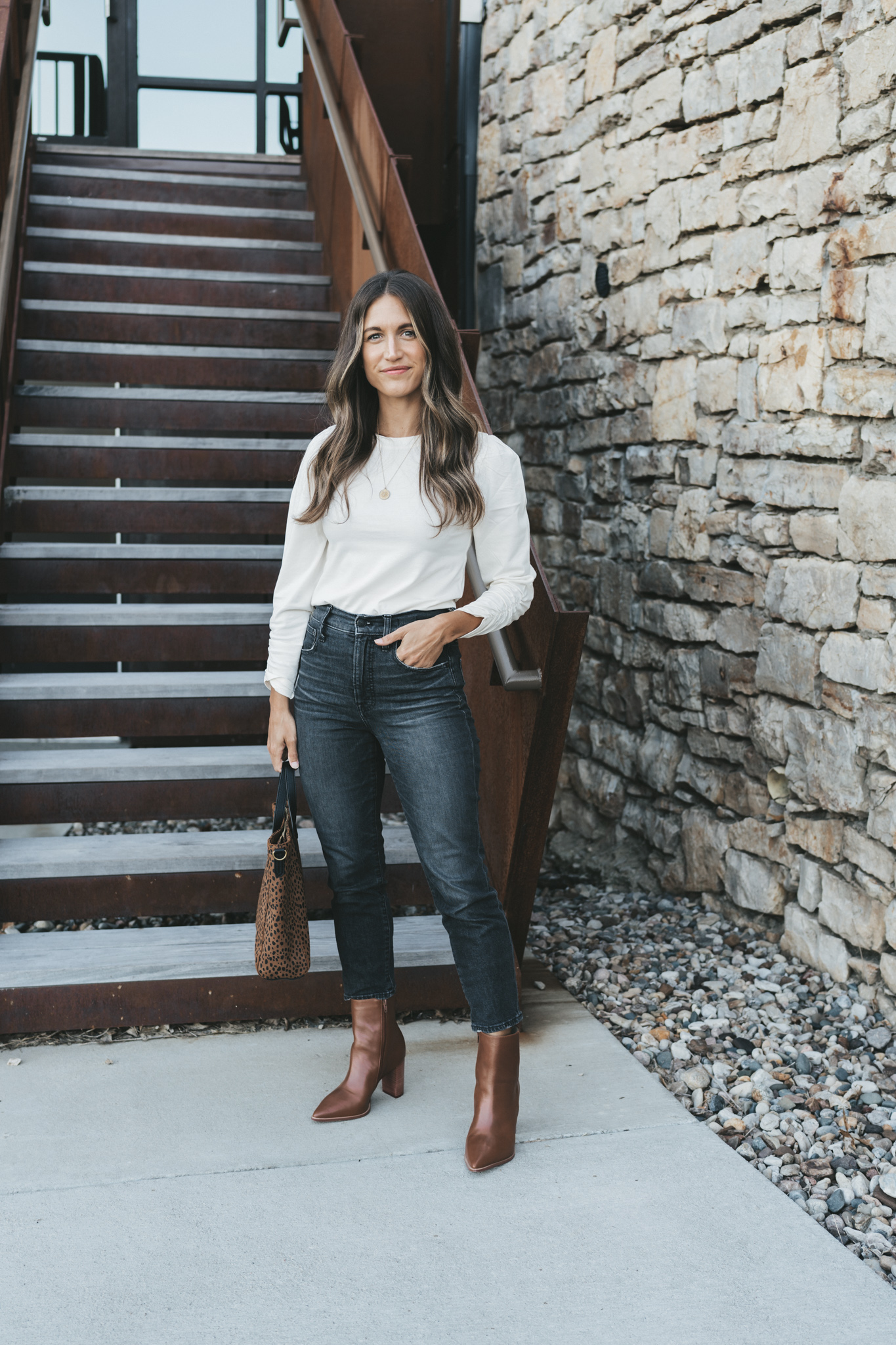 ashley wearing a white puff sleeve top, washed black denim and brown ankle boots holding a leopard print tote bag and standing outside in front of a staircase