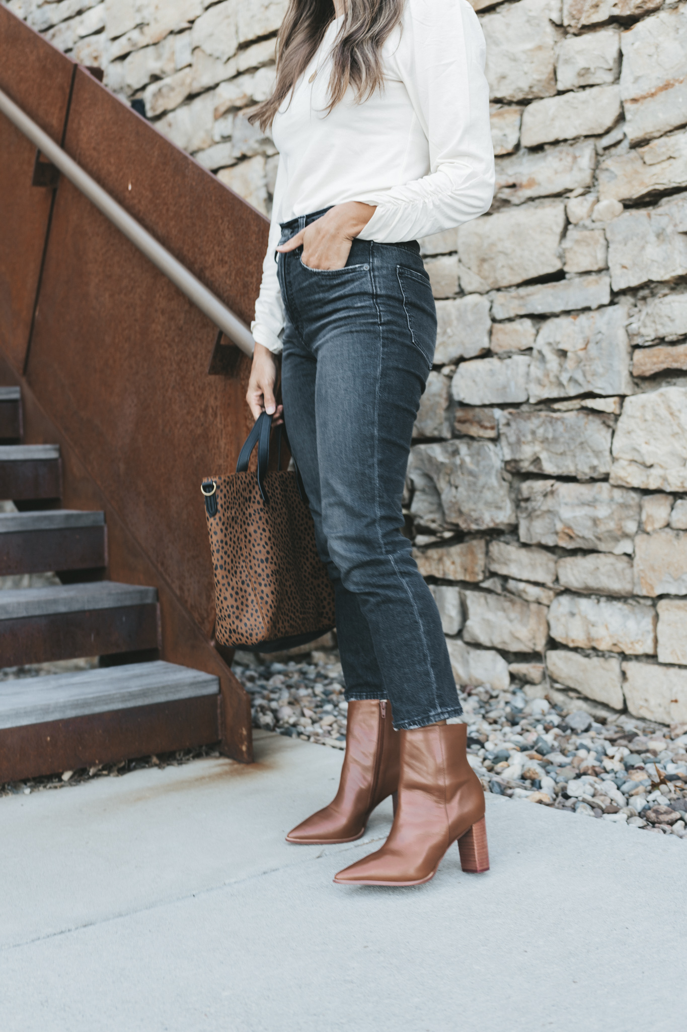 a picture of ashley wearing a white top, washed black jeans, and brown ankle boots holding a leopard print bag and standing outside next to a staircase