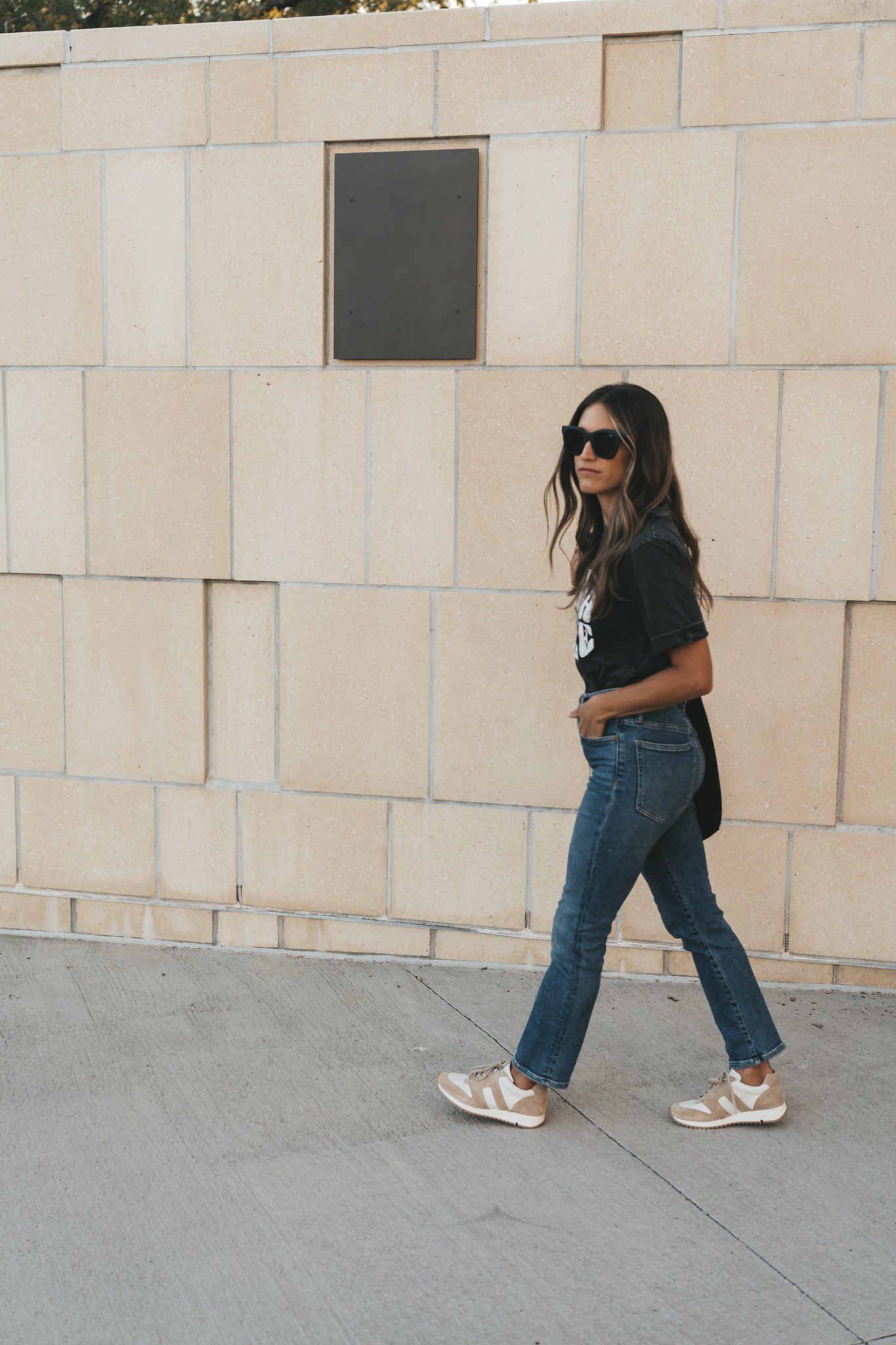 ashley wearing a black tee, medium wash jeans, and tan and white leather sneakers walking past a beige stone tile wall