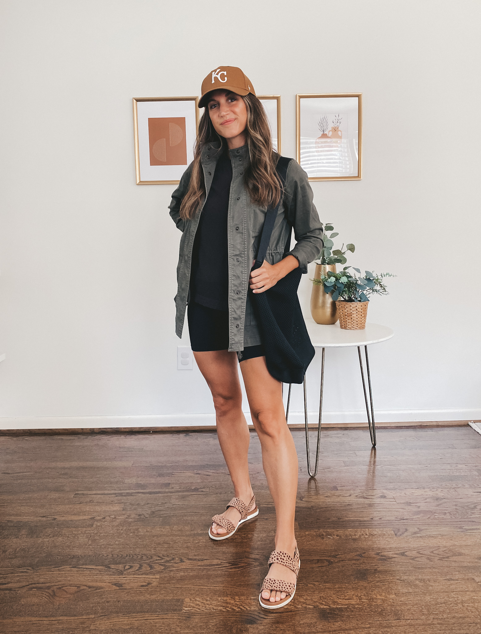 ashley wearing a green utility jacket, black tee, black biker shorts, a black tote bag and leopard print sandals with a brown KC baseball hat