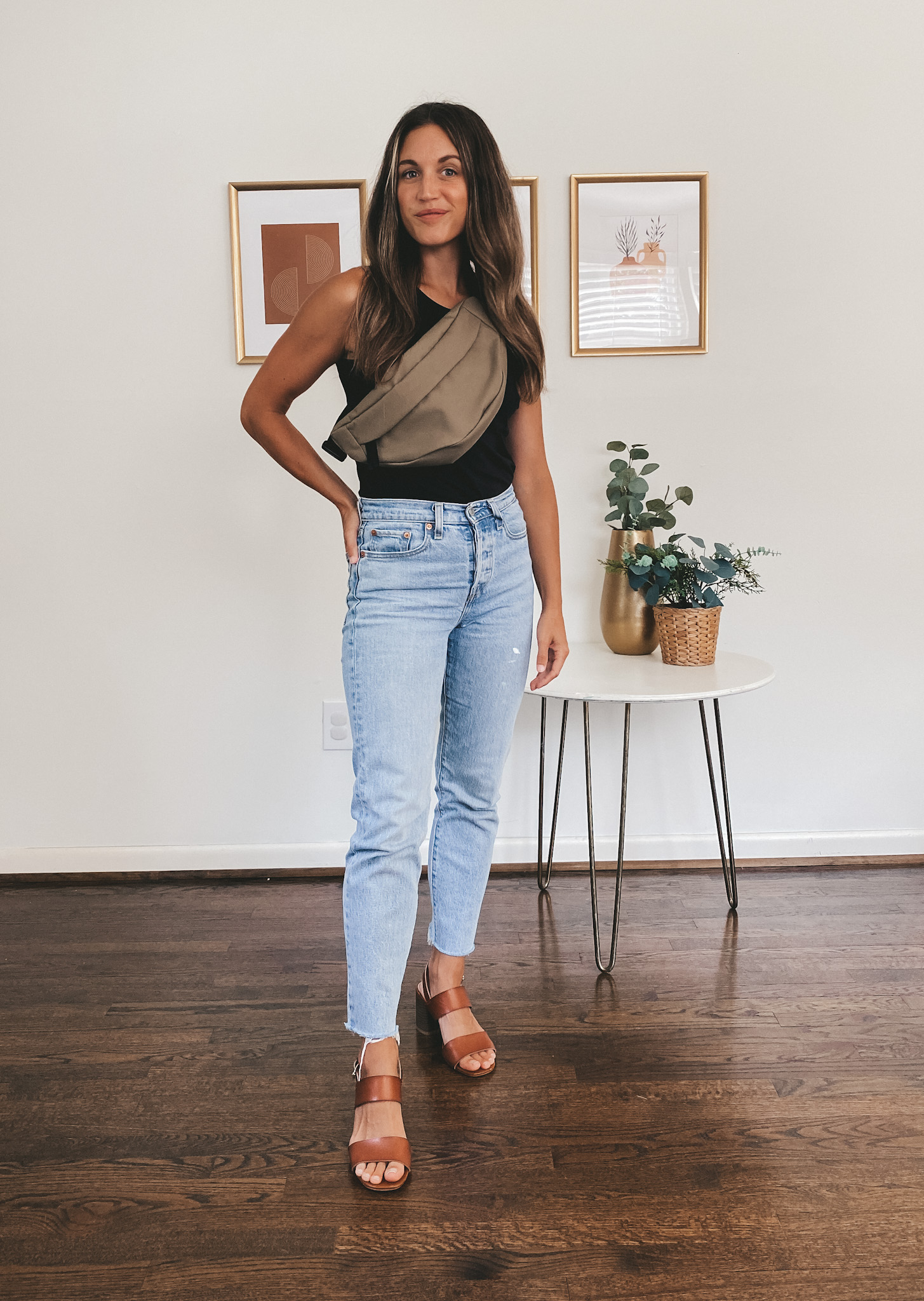 ashley wearing a black tank, light wash denim, cognac heeled sandals, and an olive green fanny pack over the shoulder