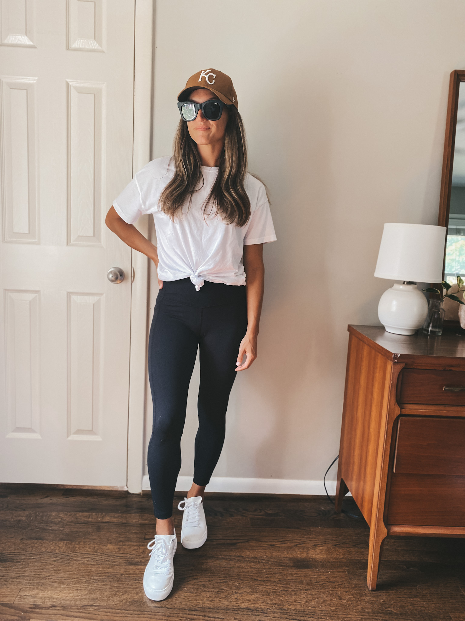 ashley wearing a white tee tied at the waist, black leggings, white sneakers, black square frame sunglasses, and a Carhartt brown KC baseball cap; early fall outfit