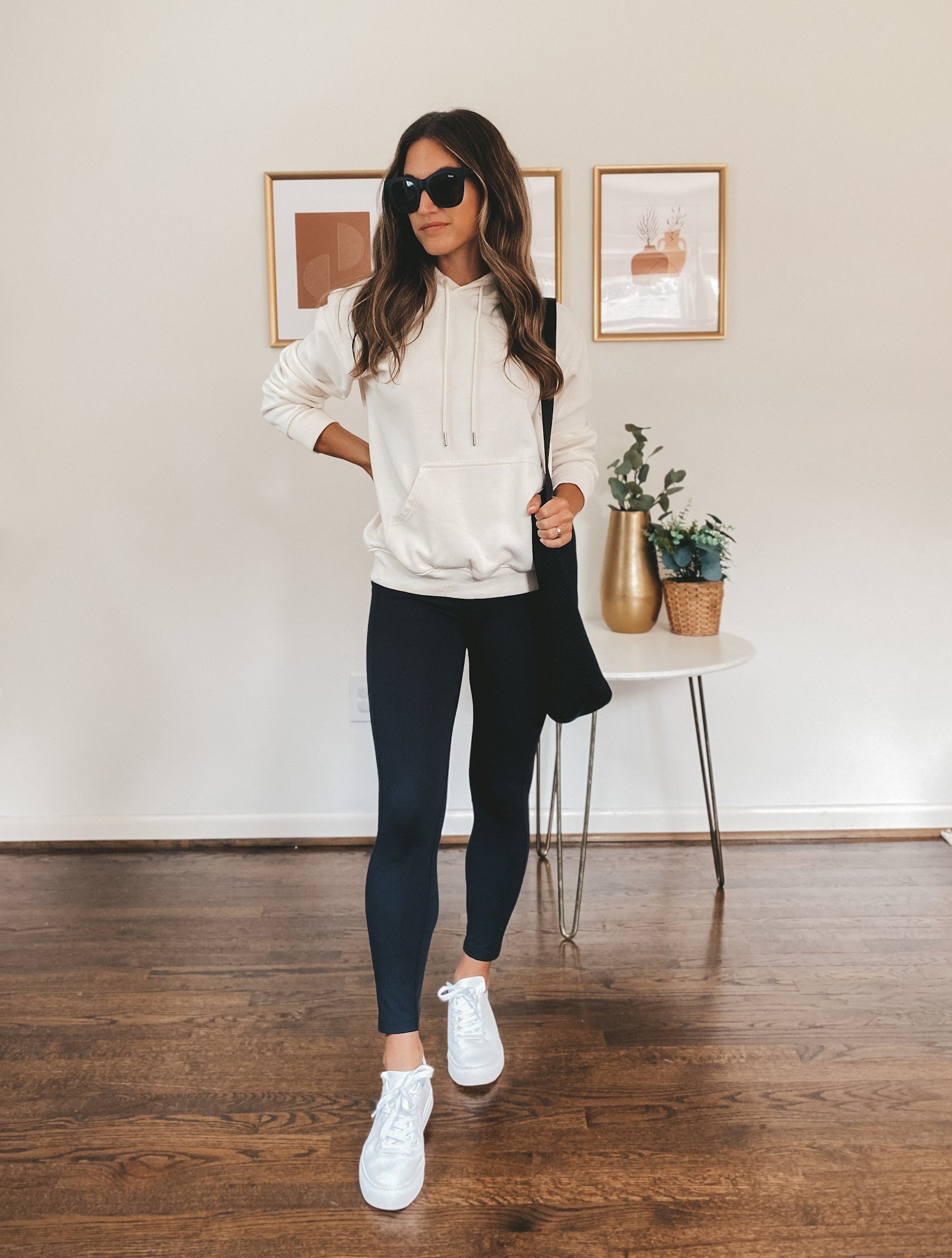 ashley wearing a cream colored hoodie, black leggings, white sneakers, a black tote bag, and black square frame sunglasses