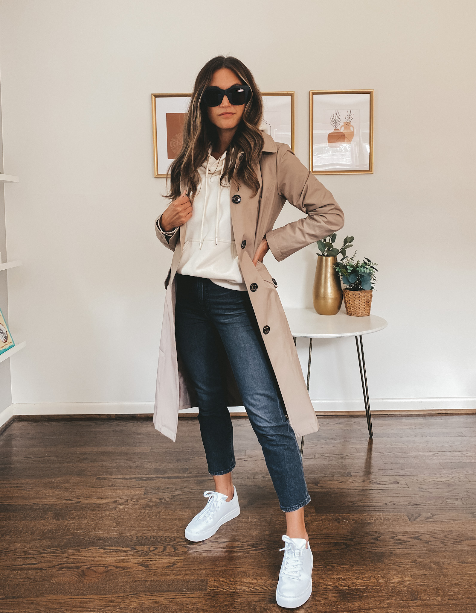 ashley wearing a cream colored hoodie, taupe trench coat, washed black jeans, white sneakers, and lack square frame sunglasses