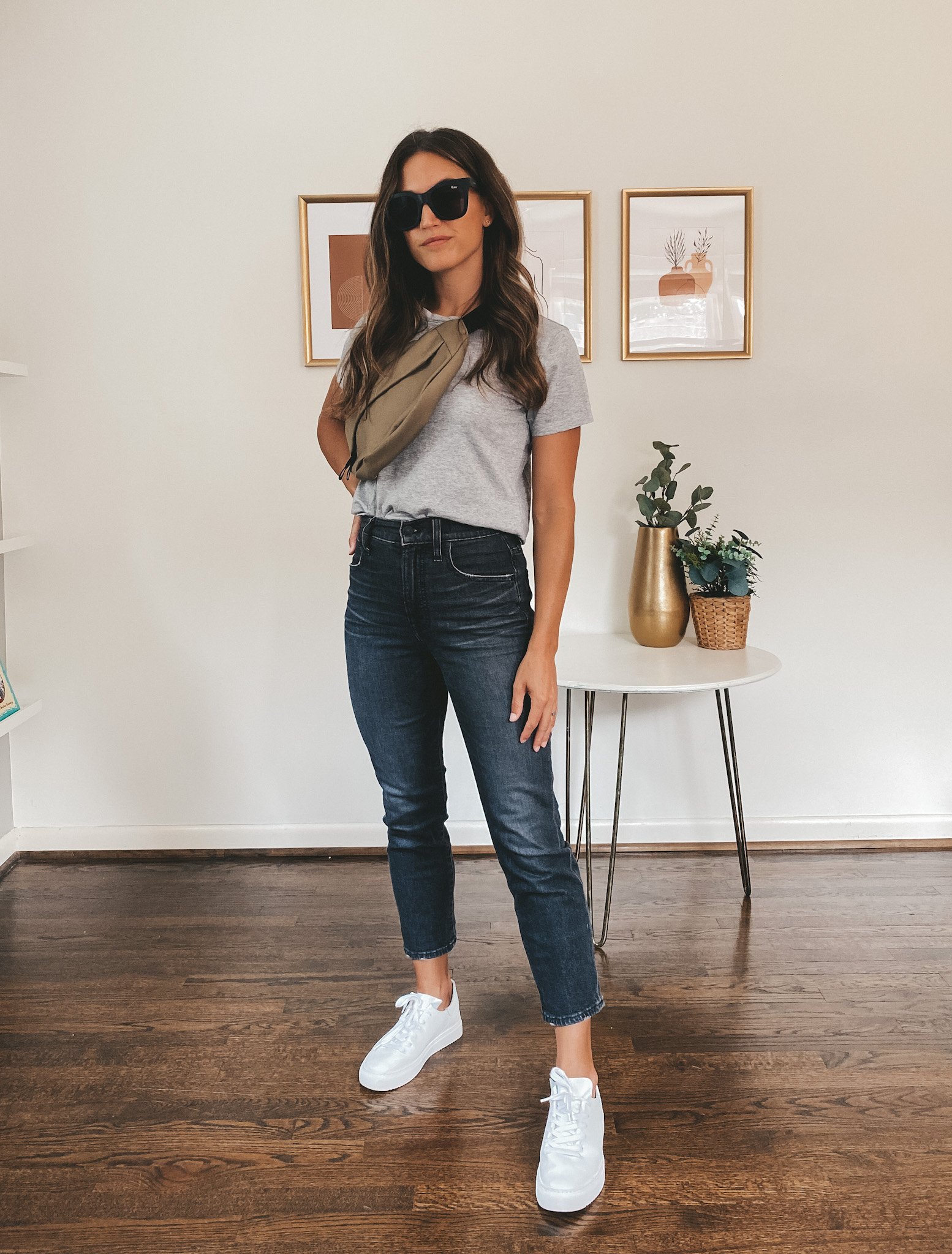ashley wearing a gray tee, washed black denim, white sneakers, an olive fanny pack, and black square frame sunglasses
