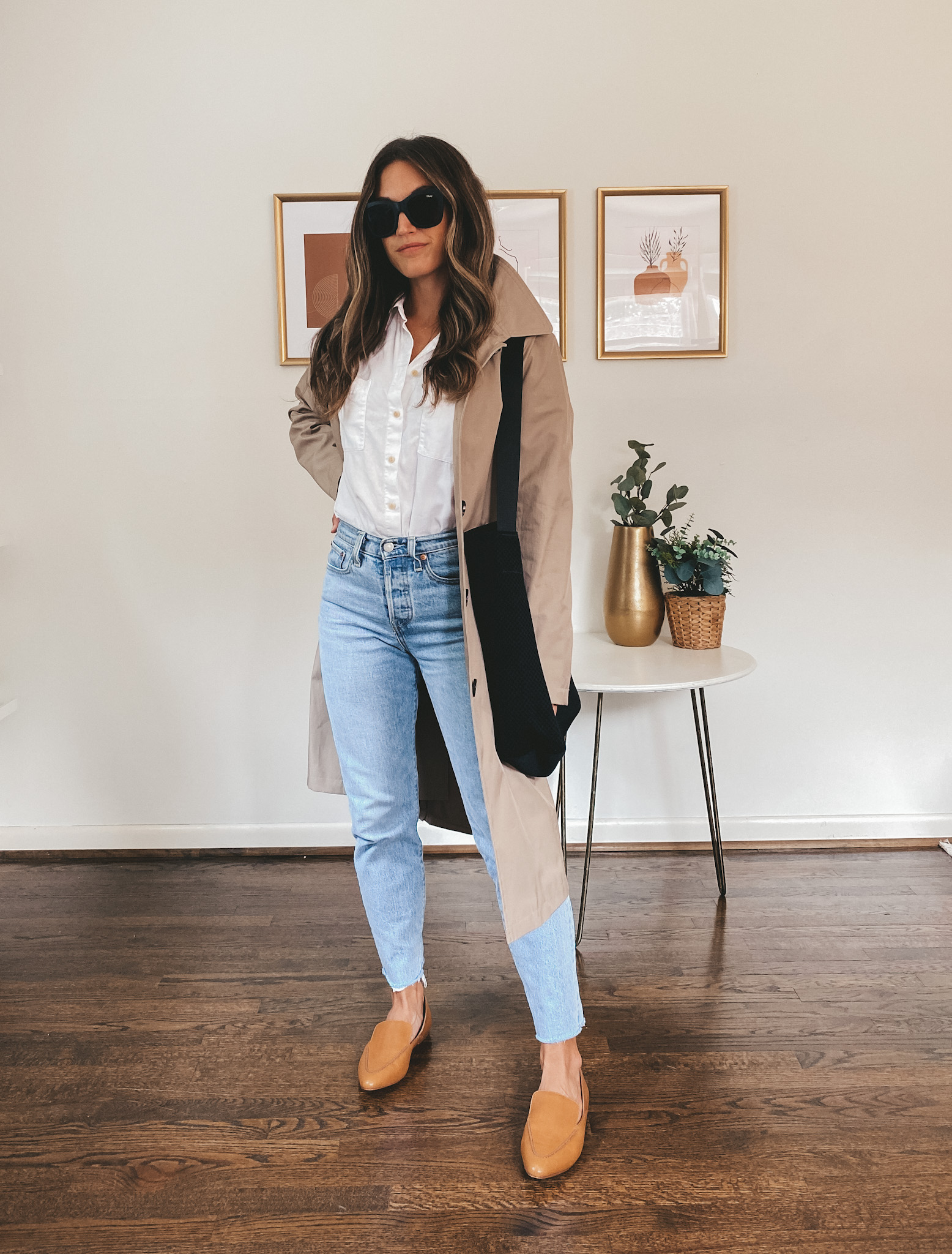 ashley wearing a white button down, taupe trench coat, light wash mom jeans, cognac loafers, a black tote bag, and square frame black sunglasses