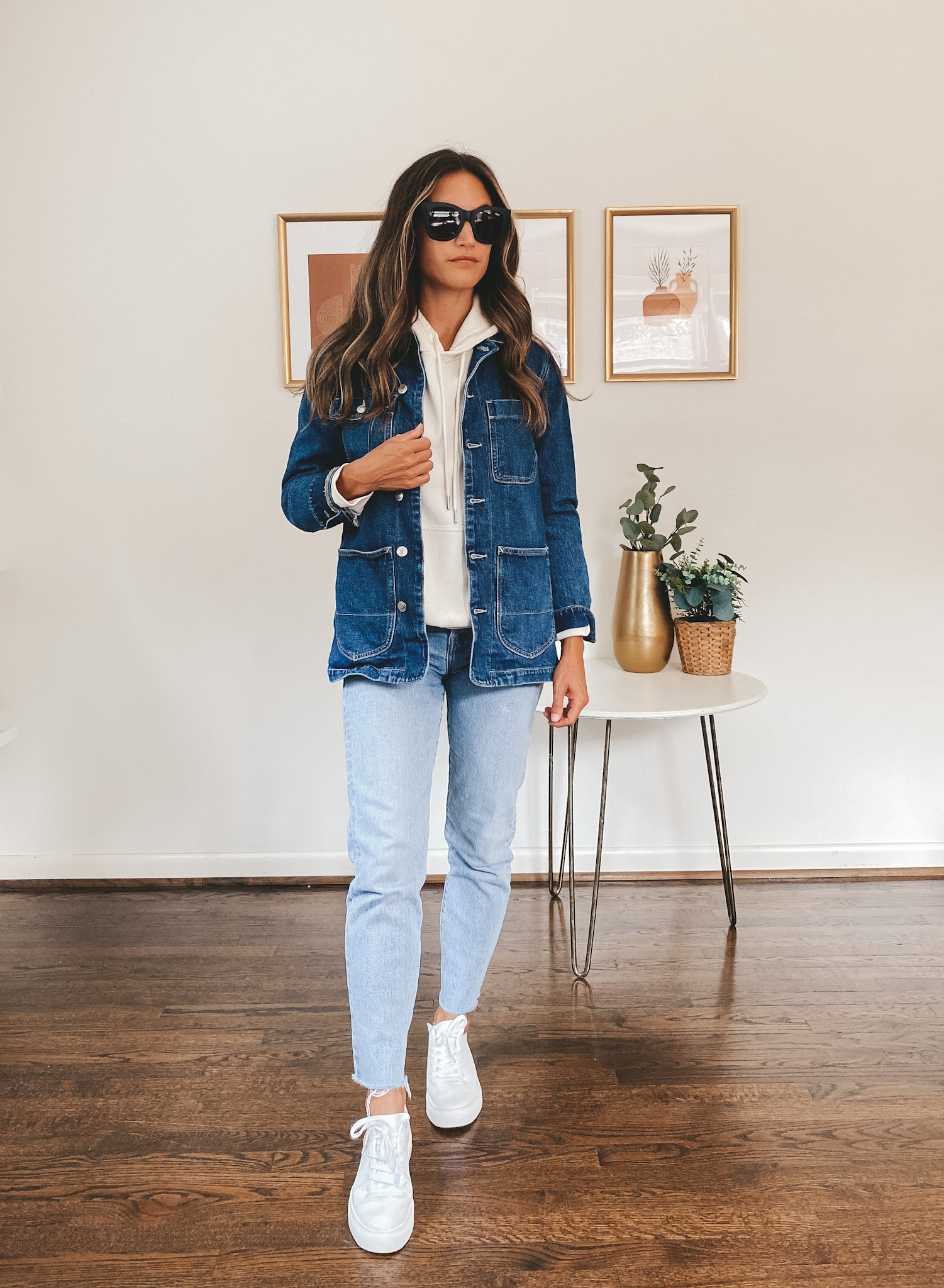ashley wearing a cream colored hoodie, denim chore jacket, light wash mom jeans, and white sneakers with black square frame sunglasses