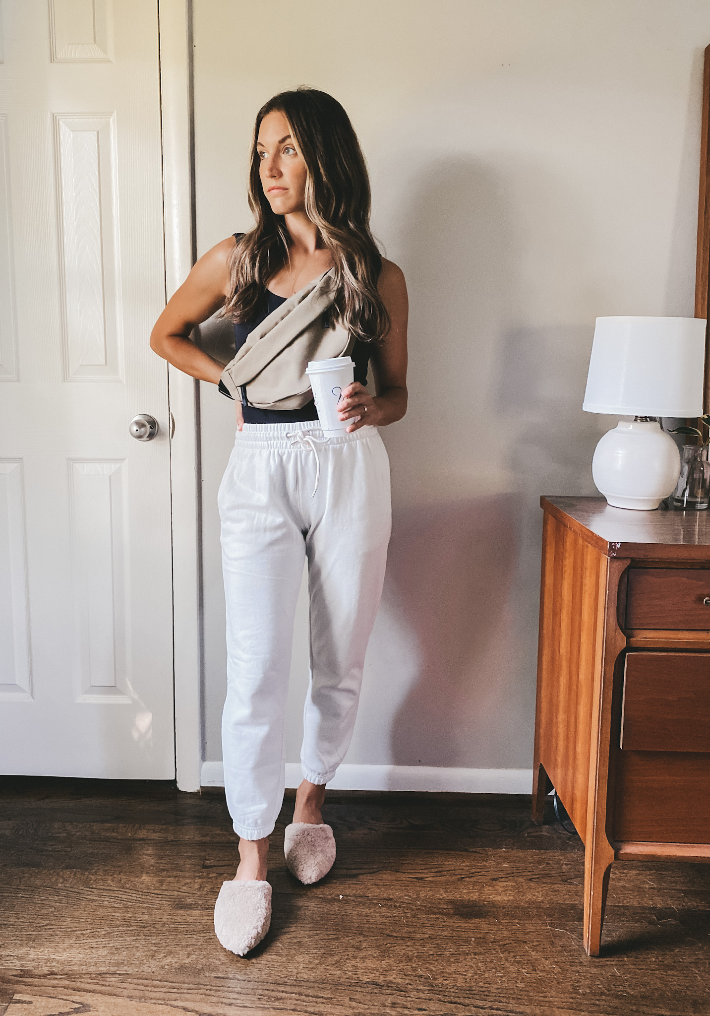 ashley wearing a black sleeveless bodysuit, white joggers, tan shearling mules, and a olive green fanny pack over the shoulder