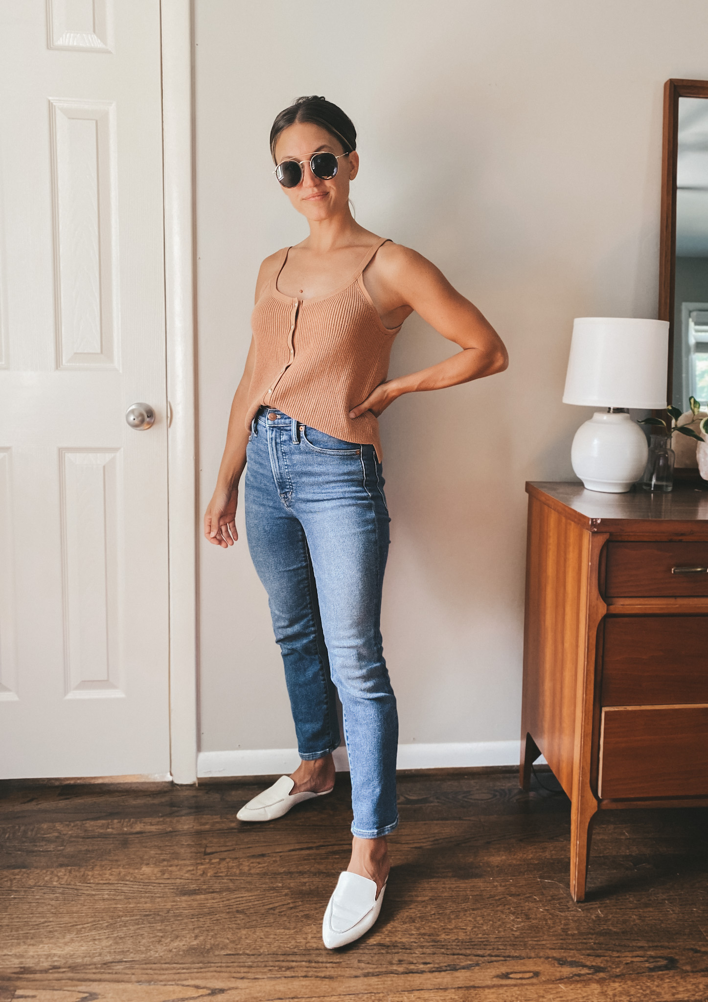 ashley wearing a tan sweater tank, medium wash denim, white leather mules and gold frame round sunglasses