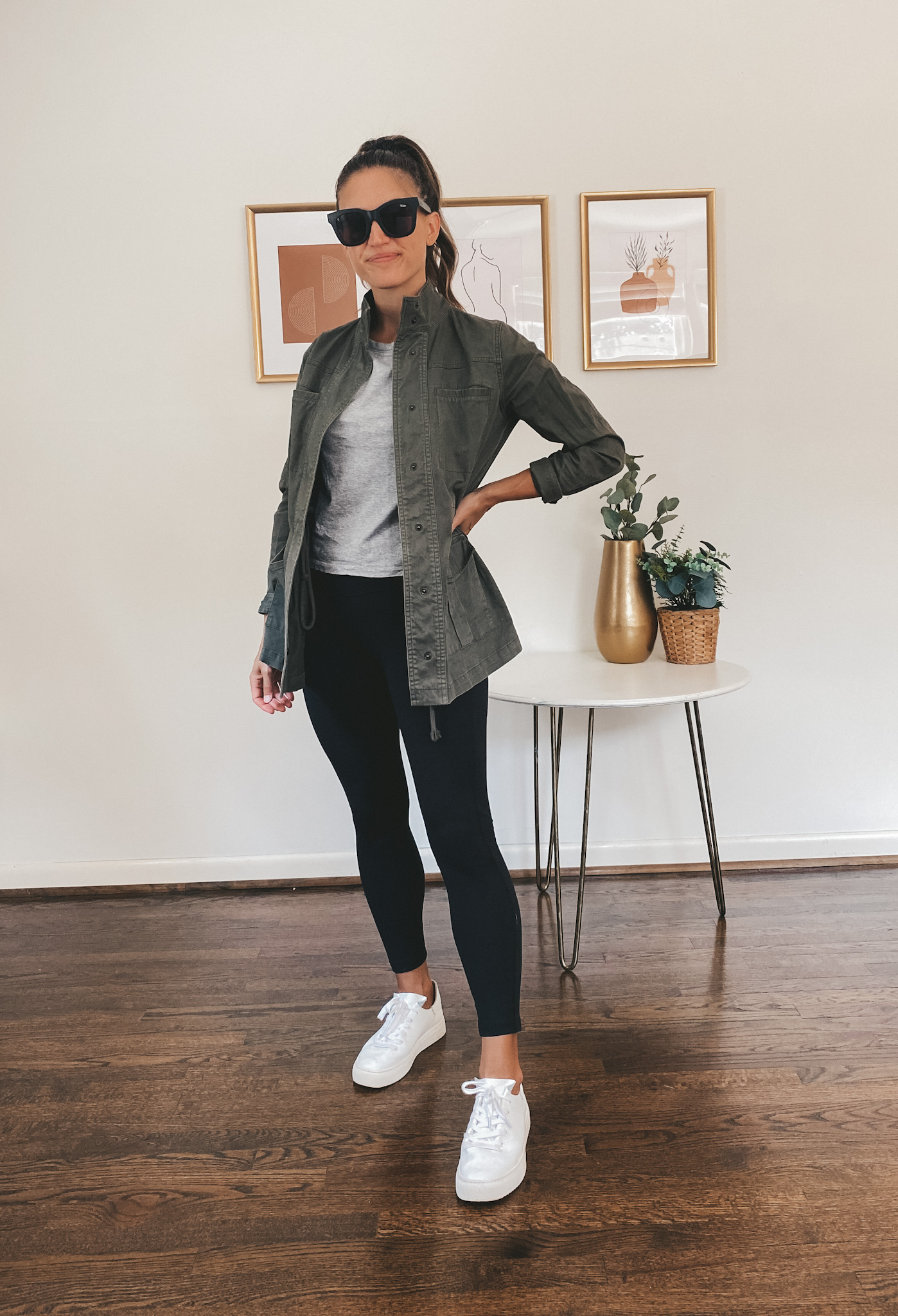 ashley wearing a gray tee, green utility jacket, black leggings, white tennis shoes, and black square frame sunglassesl
