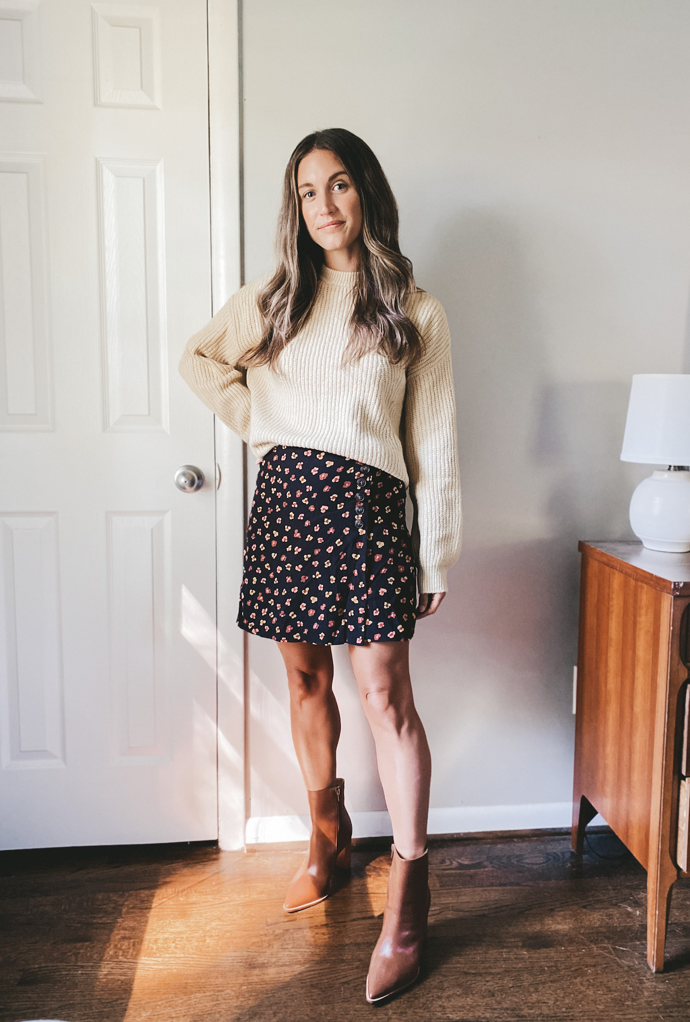 ashley wearing a cream oversized sweater, black floral mini skirt, and cognac heeled ankle boots