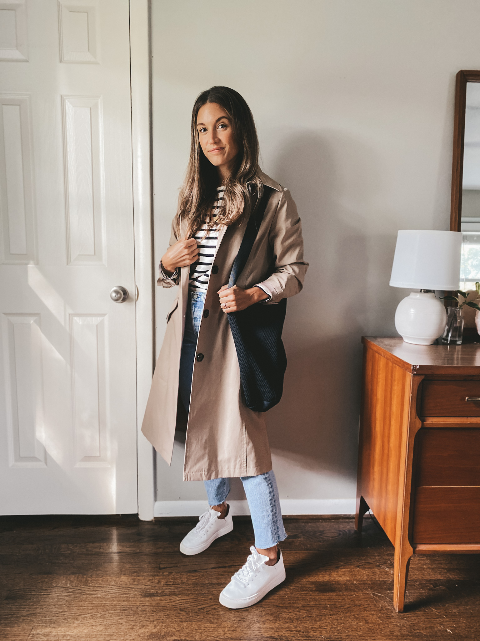 ashley wearing a striped tee, taupe trench coat, light wash jeans, white sneakers and a black tote bag