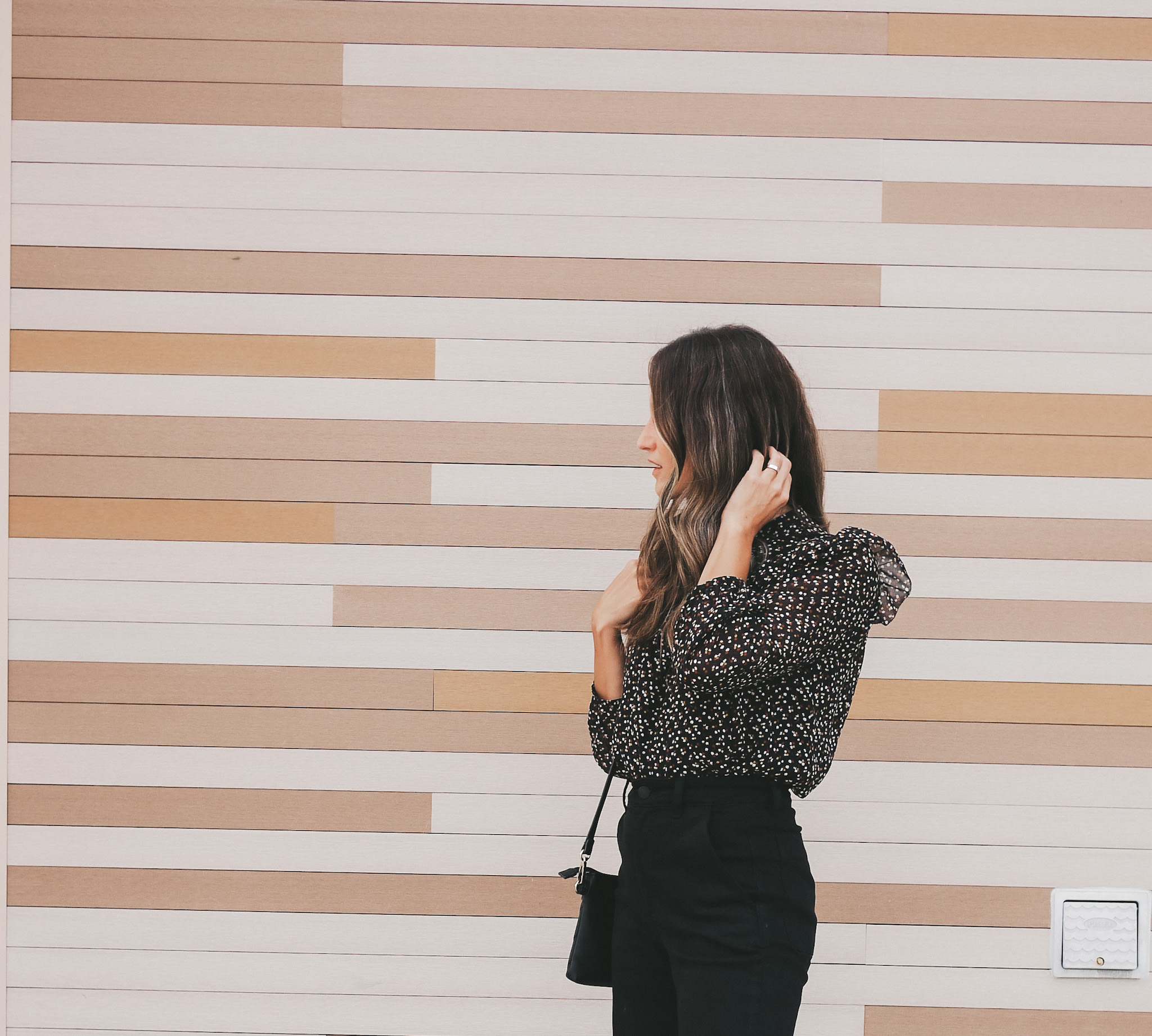 ashley standing in front of a beige, white, and brown paneled wall wearing a black floral blouse and black pants