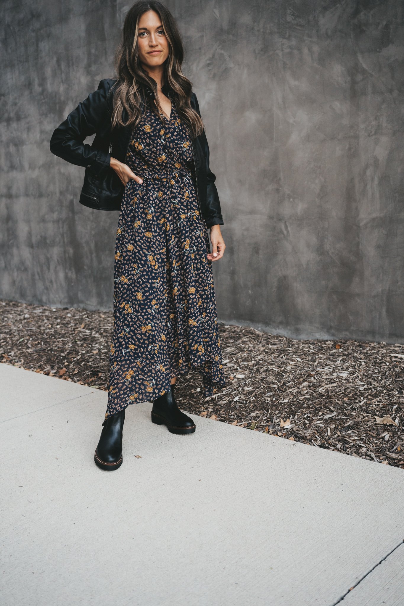 ashley wearing a black faux leather moto jacket, navy floral midi dress, and black lug sole chelsea boots