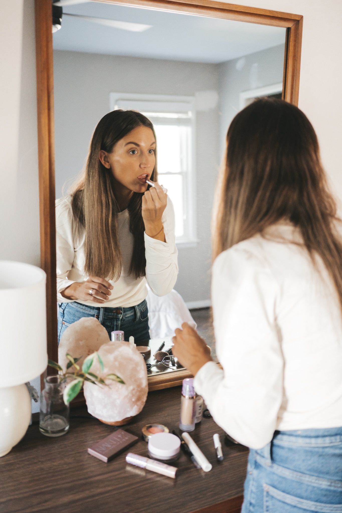 ashley looking in the mirror of a dresser applying lipstick, wearing a white top and jeans; make up is laid on the dresser