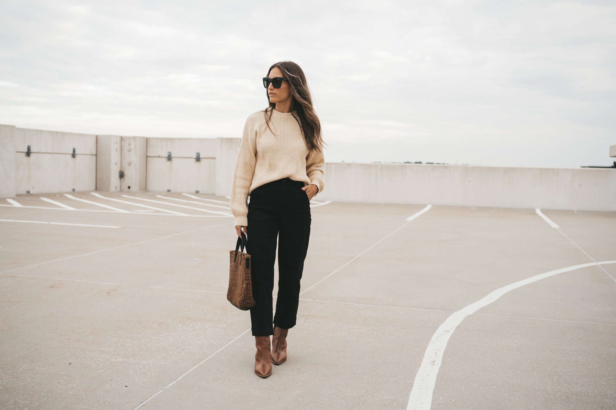 ashley wearing a cream colored sweater, black cropped pants, and cognac ankle boots with a leopard tote bag