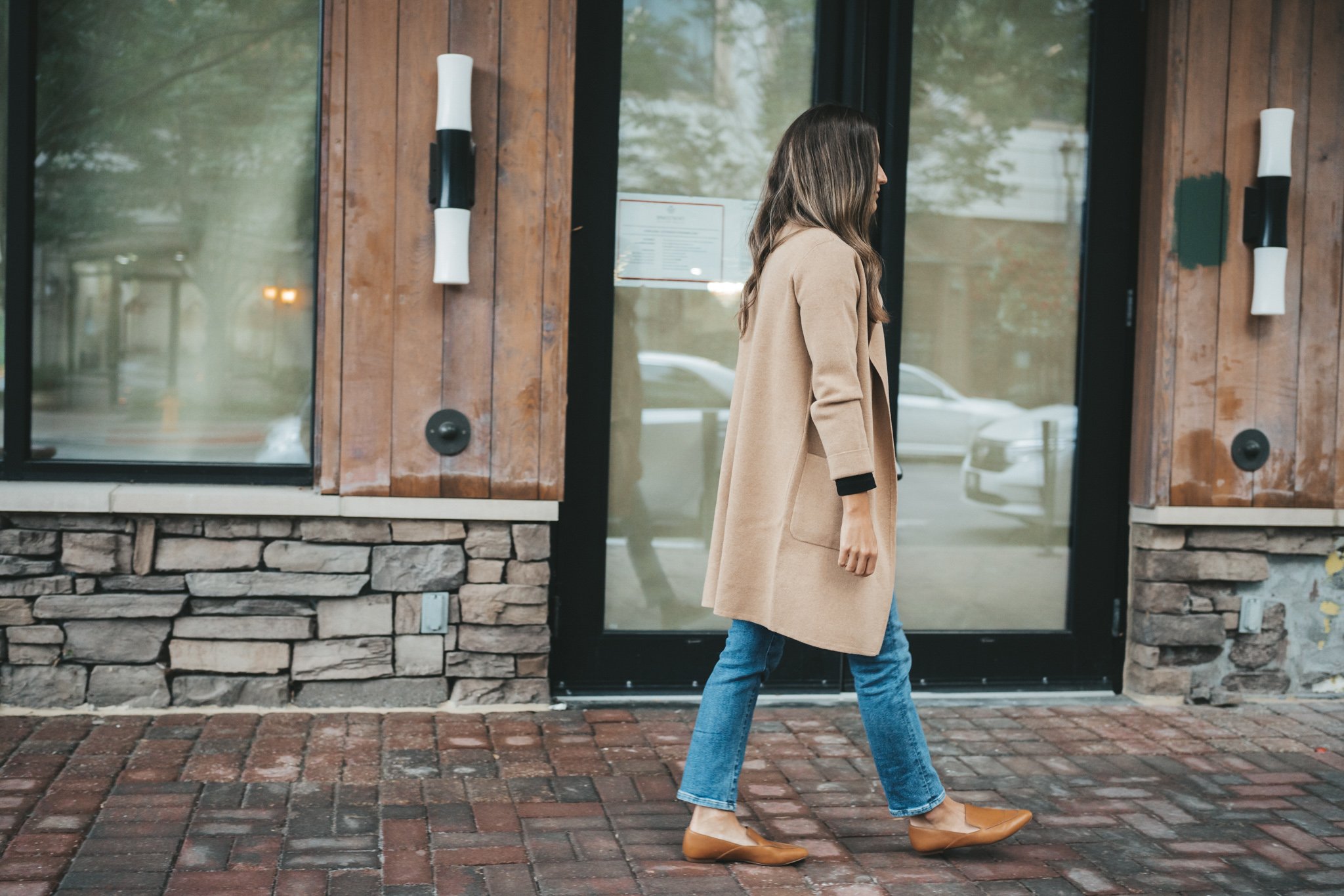 ashley wearing a tan coatigan, medium wash jeans, and cognac loafers walking down a paved sidewalk in front of a shop with stone and wood paneled walls; favorites for your fall wardrobe