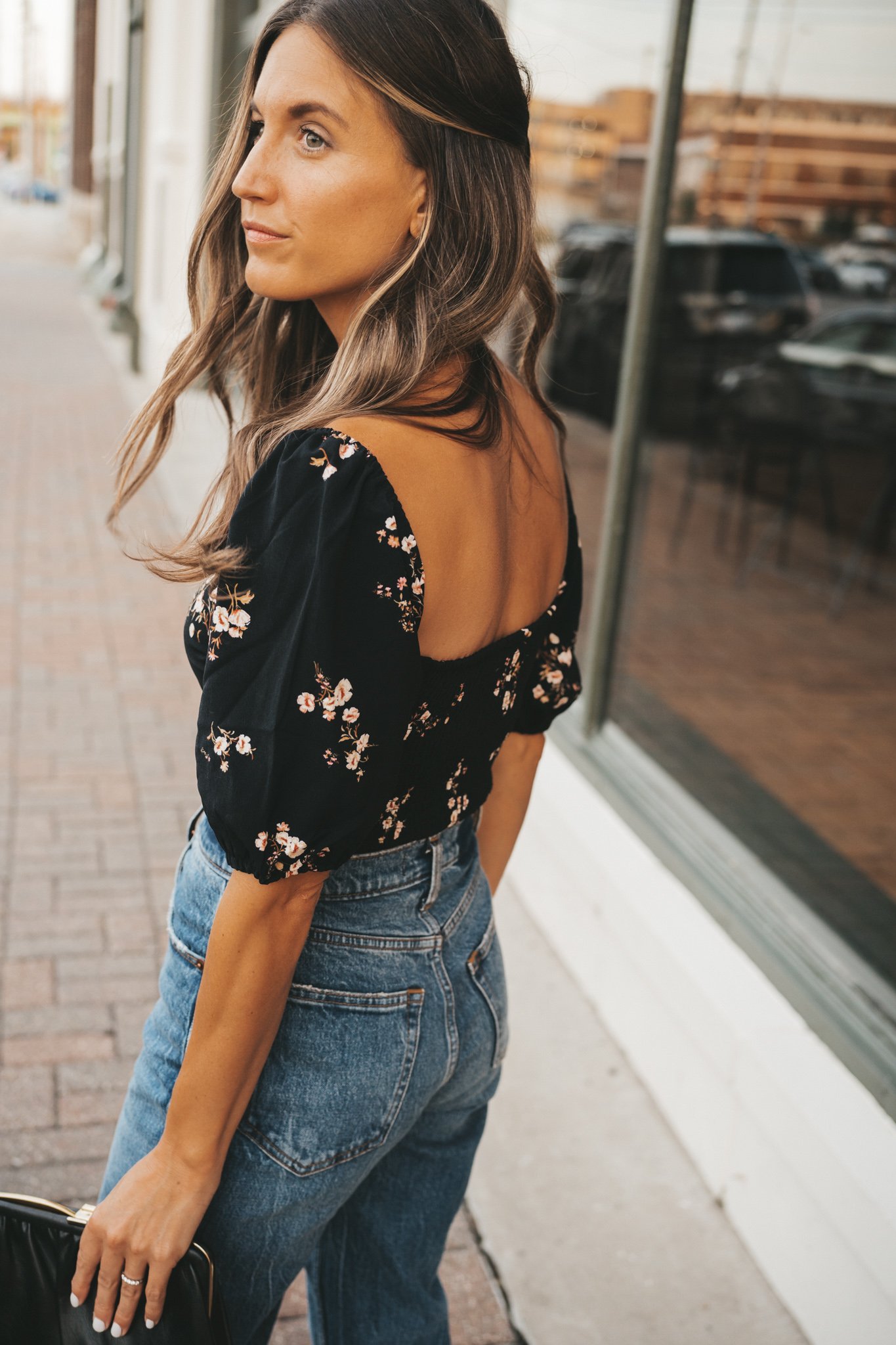 ashley looking over her shoulder wearing a black floral top and medium wash jeans standing next to the window of a shop