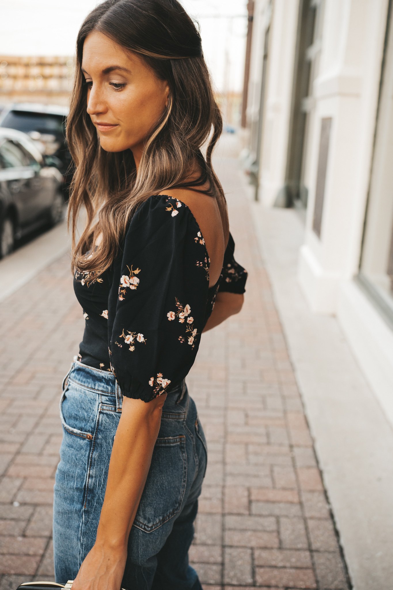 ashley wearing a date night outfit of a black floral top, medium wash jeans standing next to the window of a shop and carrying a vintage black clutch