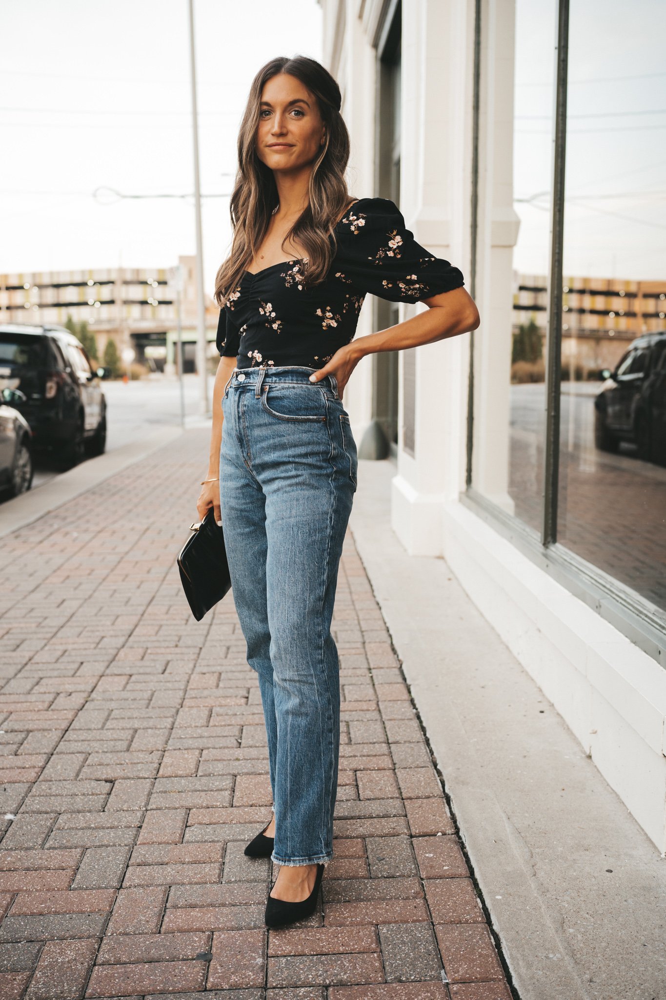 ashley wearing a fall date night outfit with a black floral top, straight leg jeans and black pumps standing on a paved sidewalk next to a shop