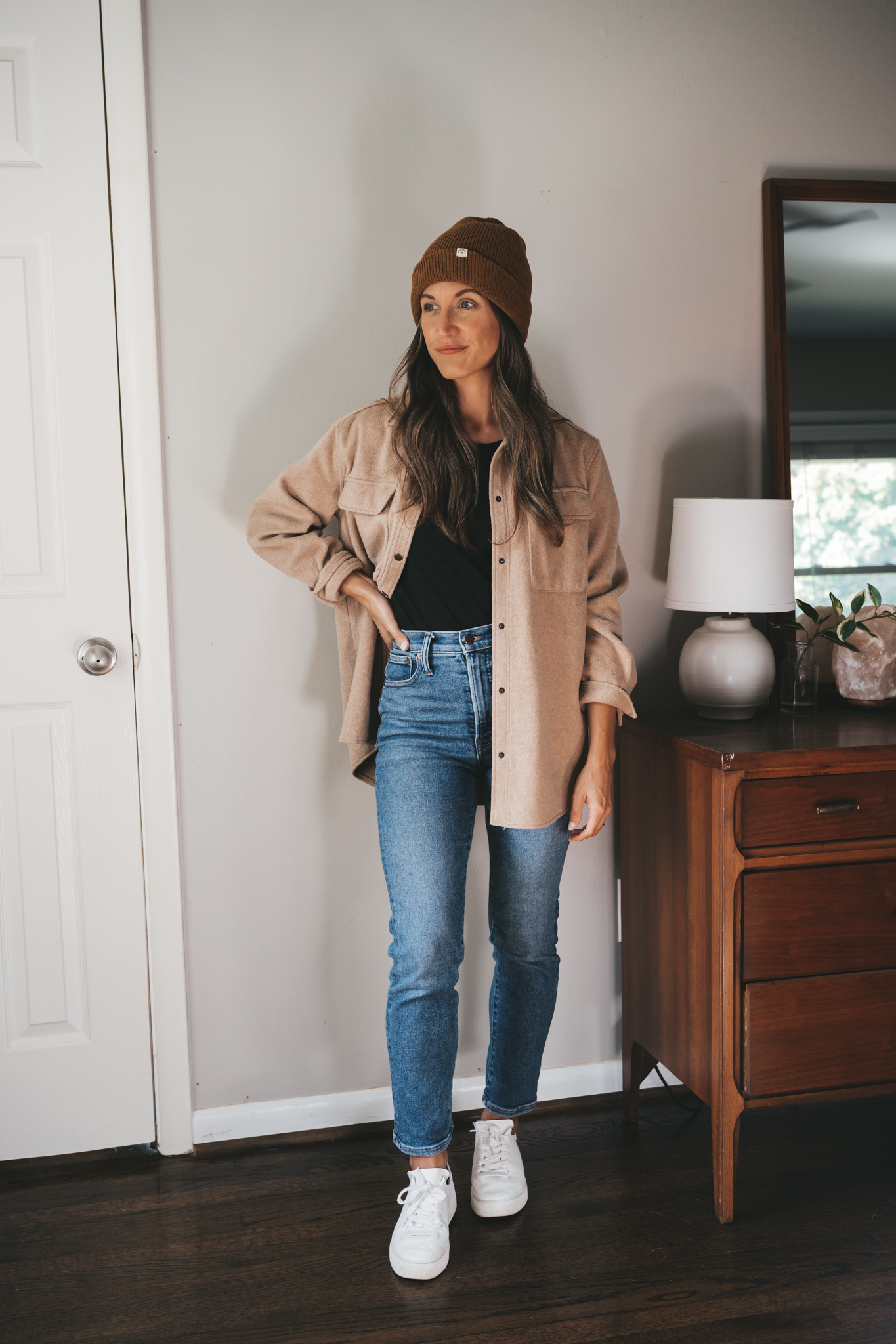 ashley wearing a black tank, tan shacket, medium wash jeans, white sneakers and a cedar colored beanie