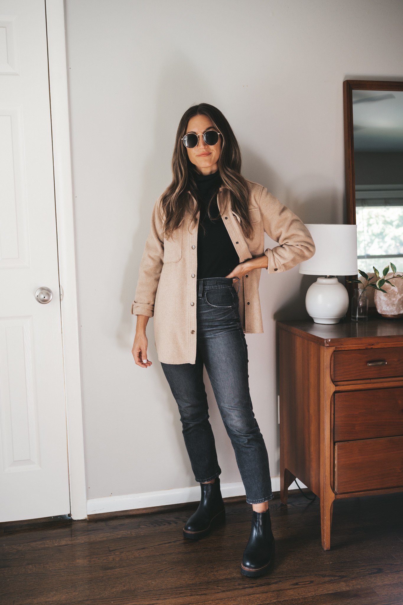 ashley wearing a black turtleneck, tan shacket, washed black jeans, black Chelsea boots, and round gold frame sunglasses