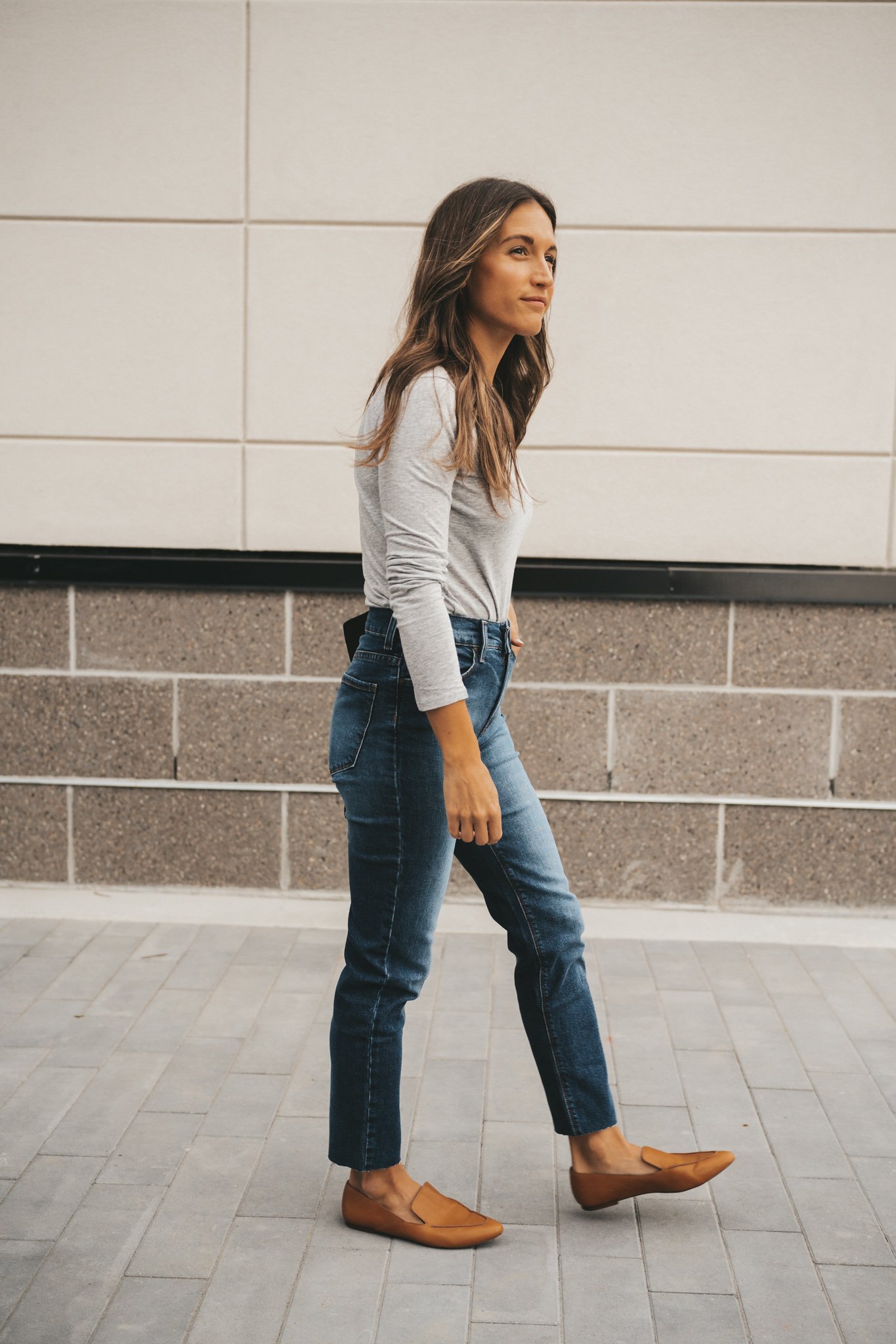 ashley wearing a long-sleeved gray crew neck tee from Mott and Bow, medium wash boyfriend denim and cognac loafers walking next to a gray and white building