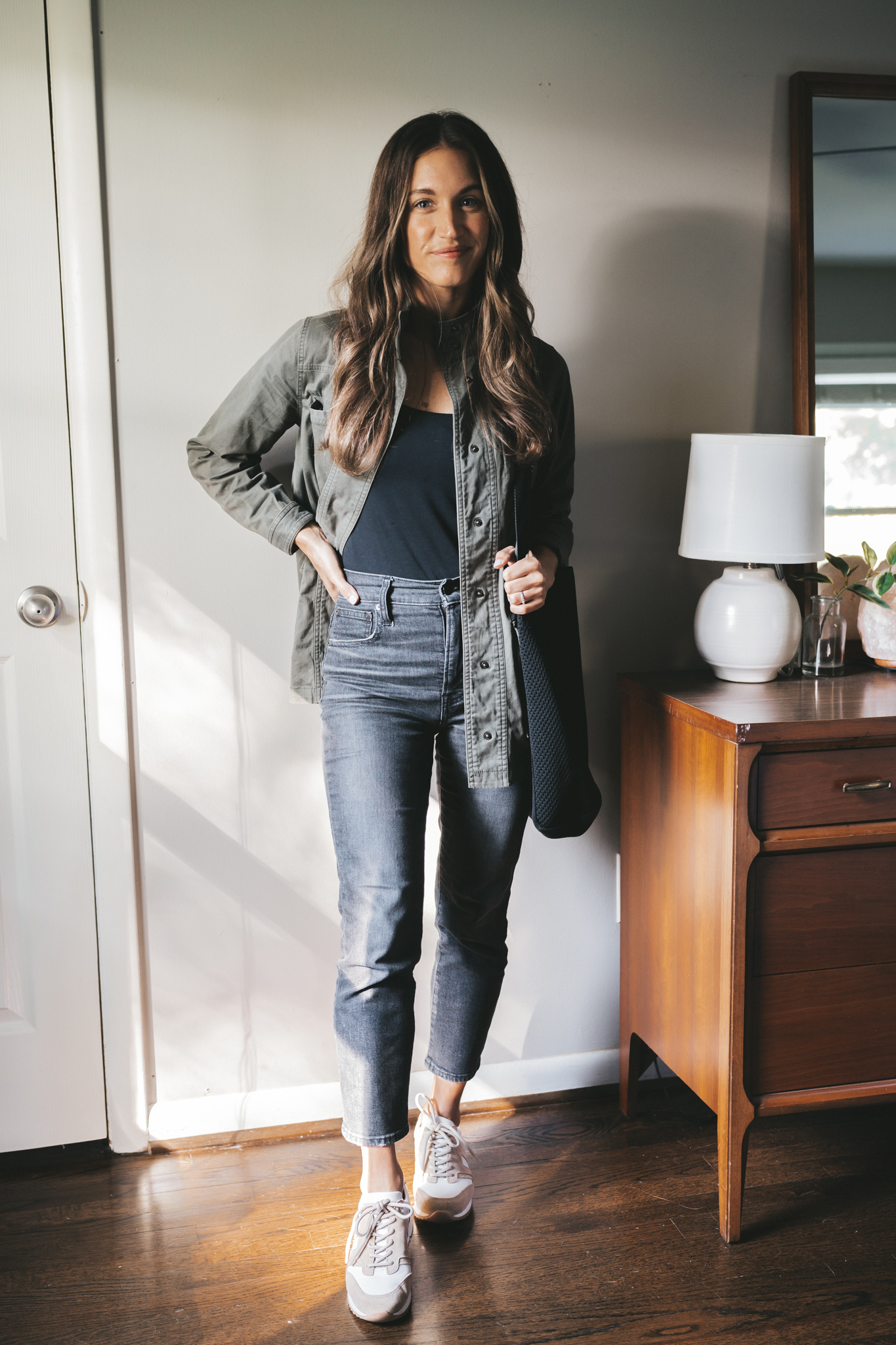 ashley wearing a black bodysuit, green utility jacket, washed black jeans, white and tan leather sneakers and a black tote bag