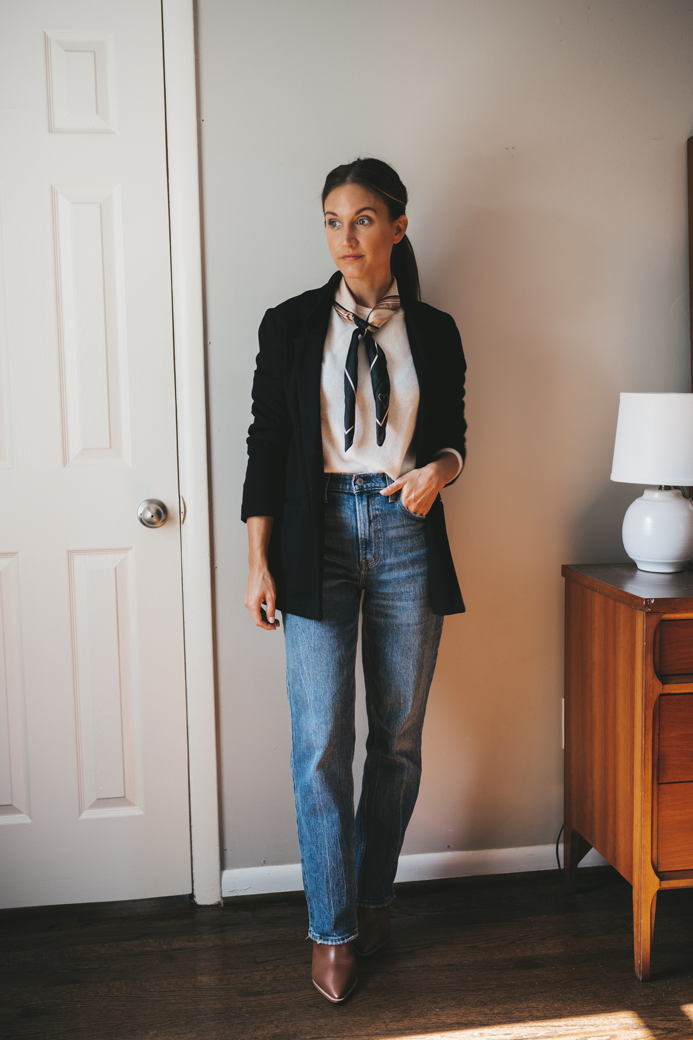 ashley wearing a cream colored cashmere sweater, black blazer, patterned scarf tied at the neck, straight leg medium wash denim, and heeled brown ankle boots