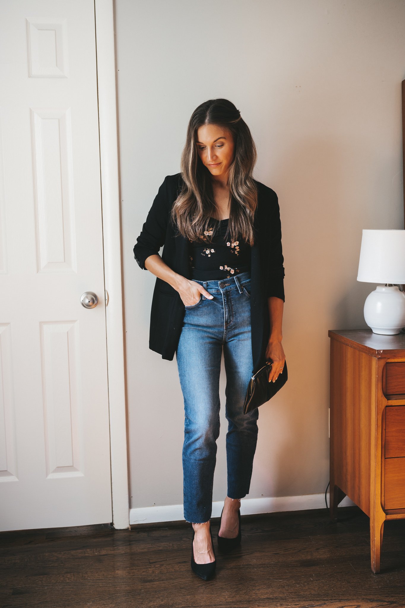 ashley wearing a black floral top, black blazer, dark wash denim, and black pumps with a black vintage clutch