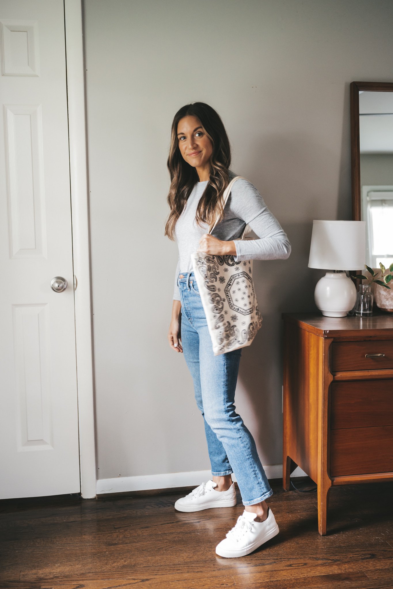 ashley wearing a long-sleeved gray tee, medium wash mom jeans, white sneakers, and a patterned canvas tote bag