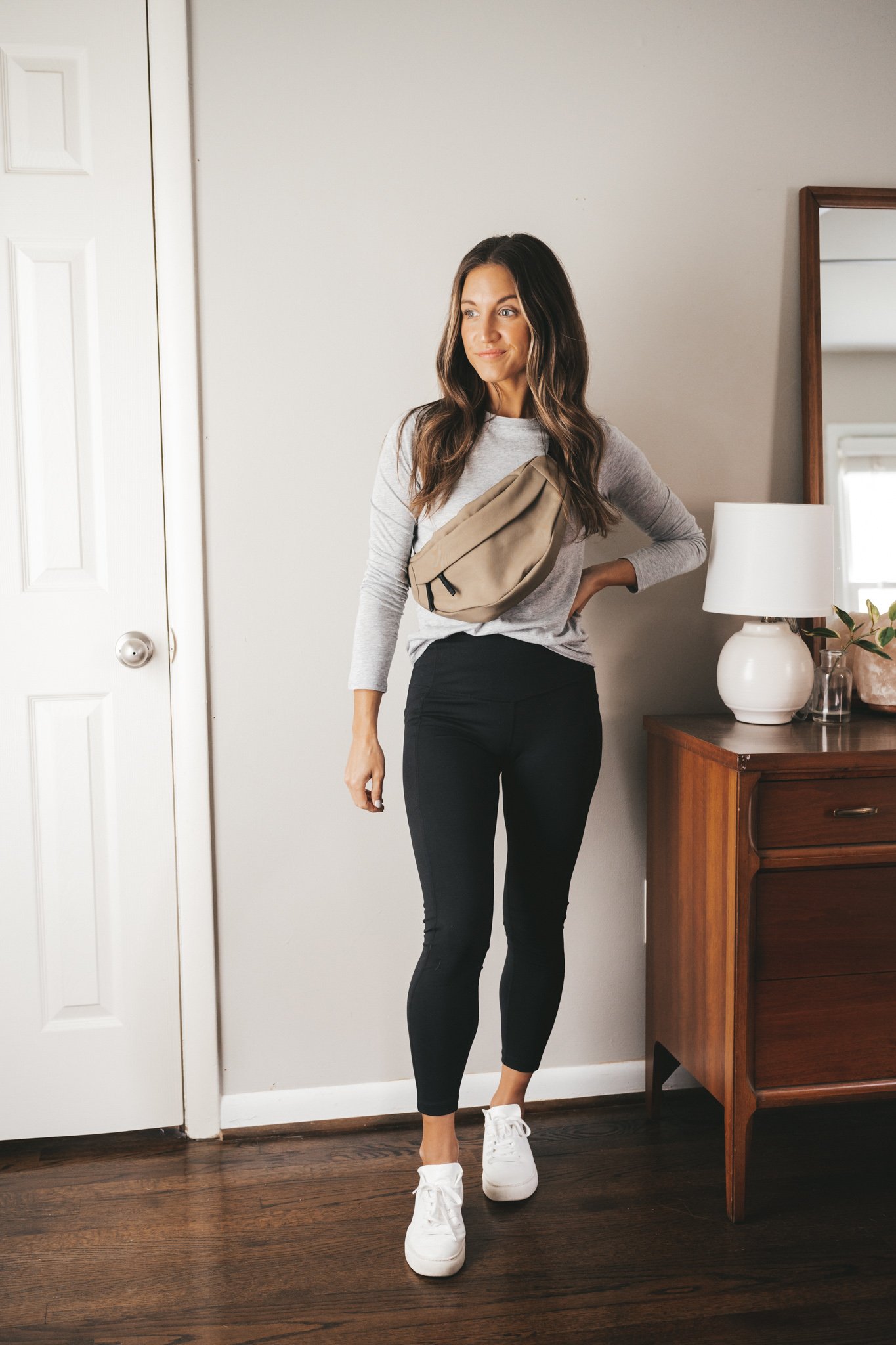 ashley wearing a gray long-sleeved tee, black leggings, white sneakers, and an olive green belt bag worn over the shoulder