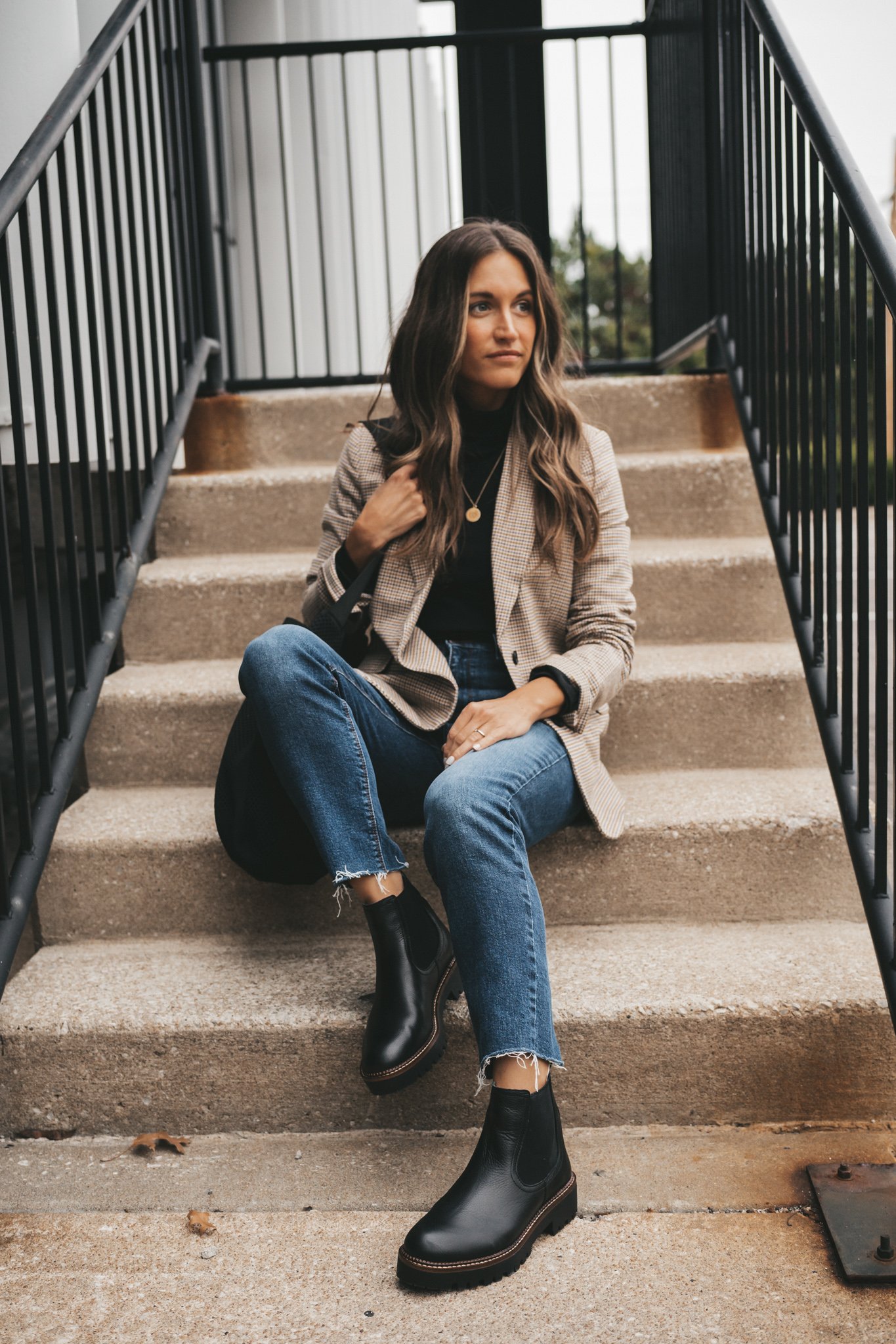 ashley wearing a black turtleneck under a plaid blazer, medium wash mom jeans, and black lug sole boots sitting on cement stairs with a black metal railing
