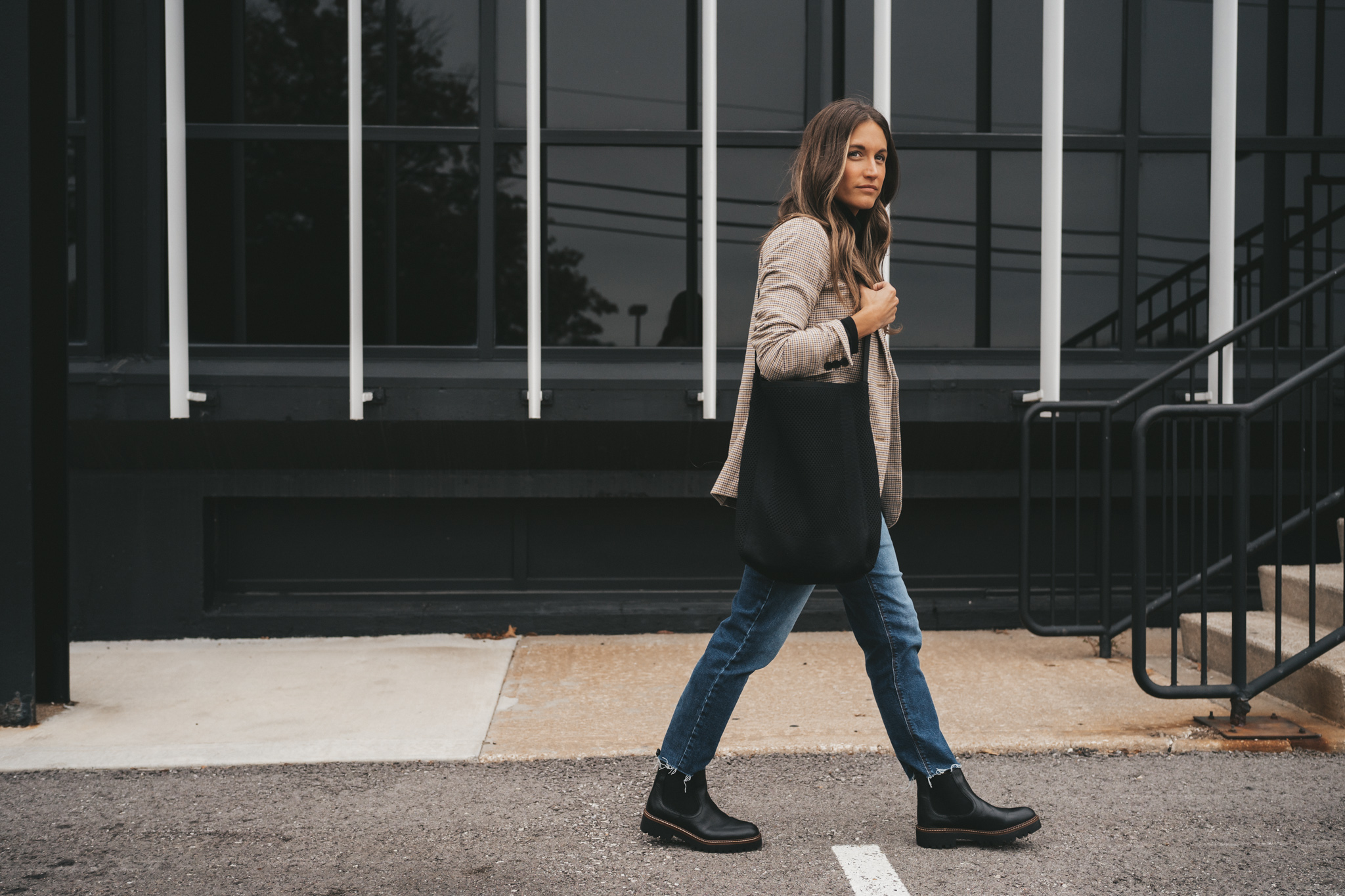 ashley wearing a black turtleneck under a plaid blazer, medium wash mom jeans, and black lug sole boots walking in front of a black building with large windows