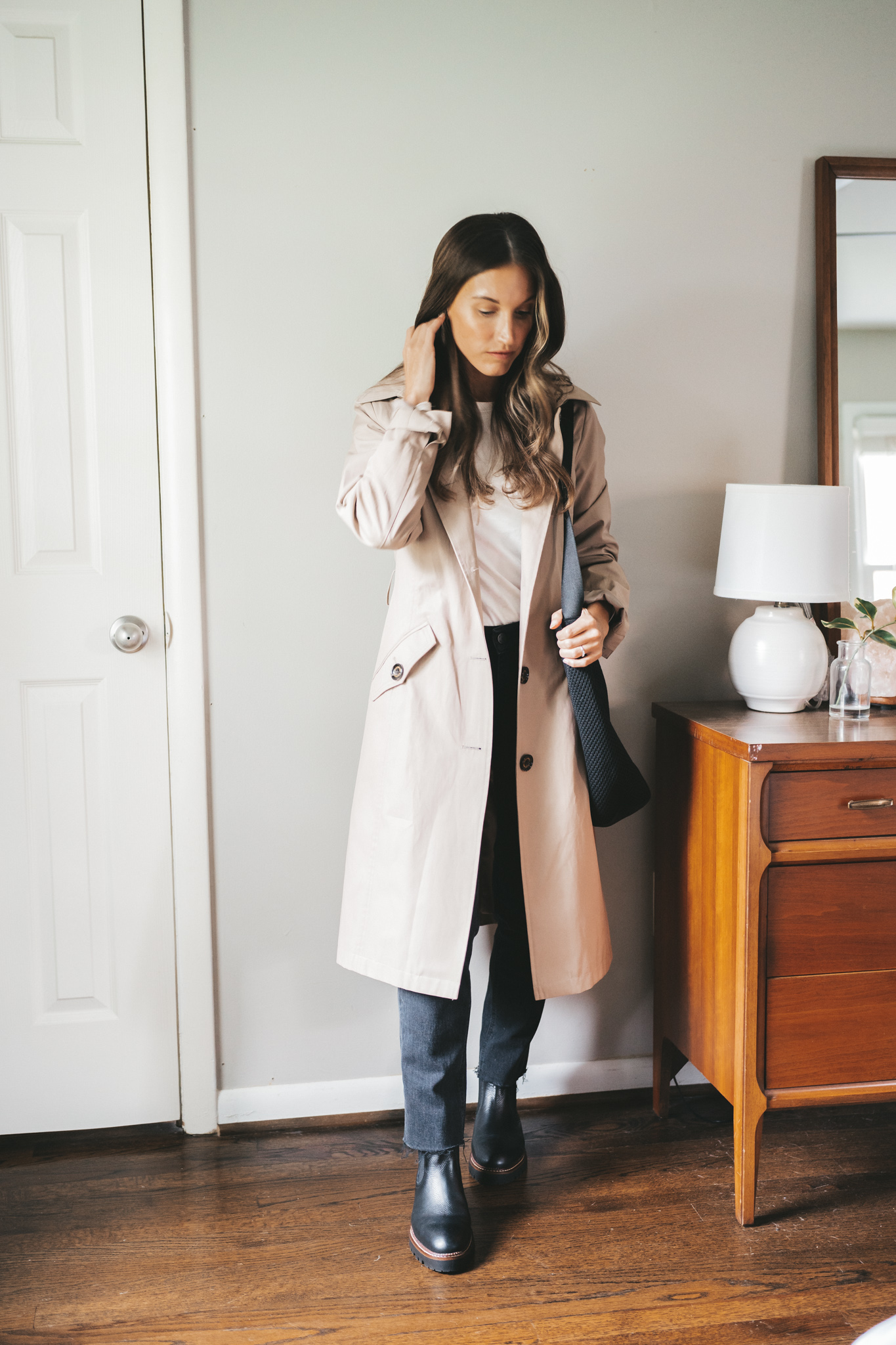 ashley wearing a white top under a taupe trench coat, black boyfriend jeans, and black lug sole chelsea boots and a black tote bag