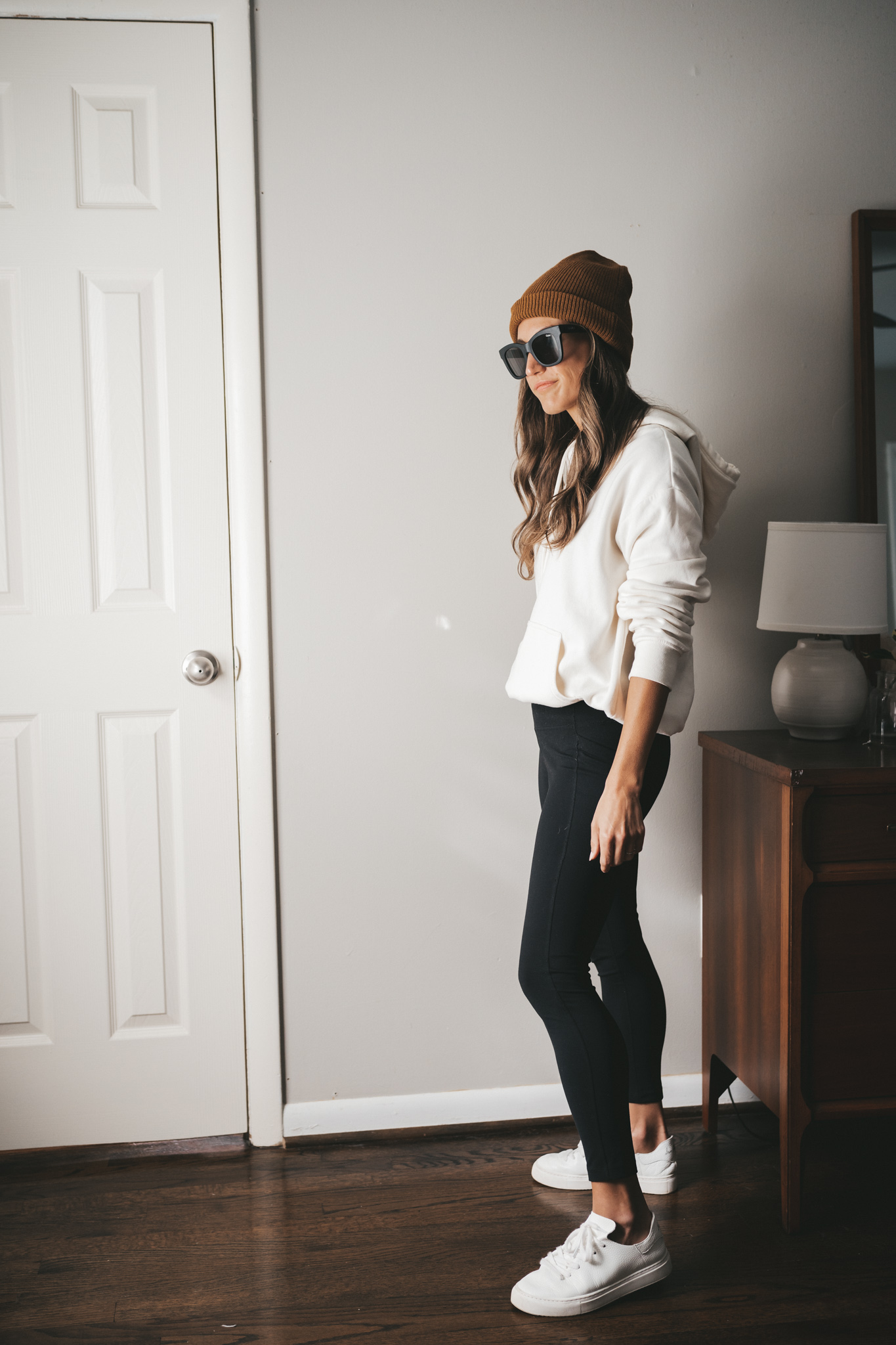 ashley wearing a cream colored hoodie, black leggings, white sneakers, black square frame sunglasses and a brown beanie