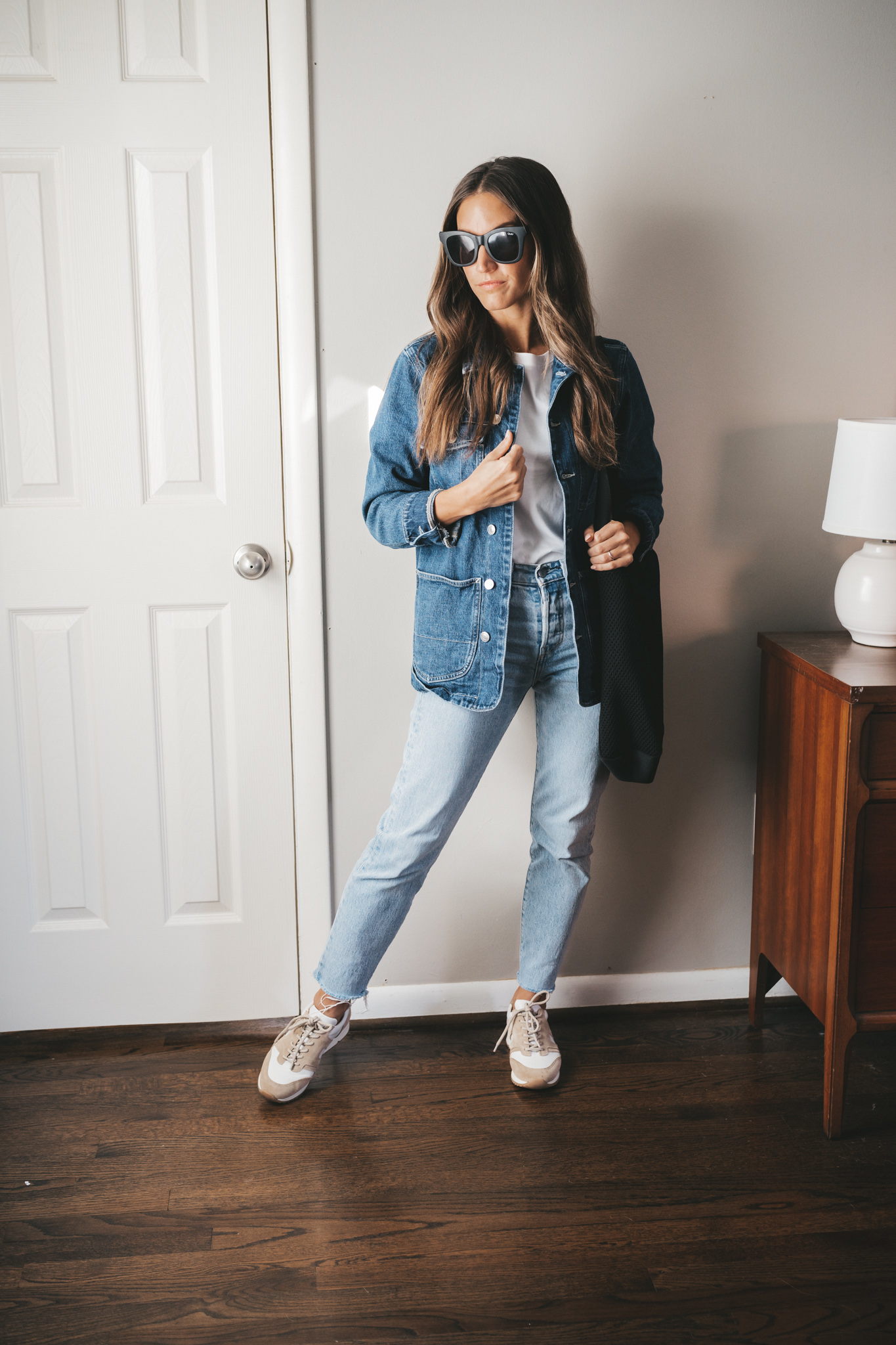 ashley wearing a white tee under a denim chore jacket, light wash mom jeans, white and tan leather sneakers, and black square frame sunglasses