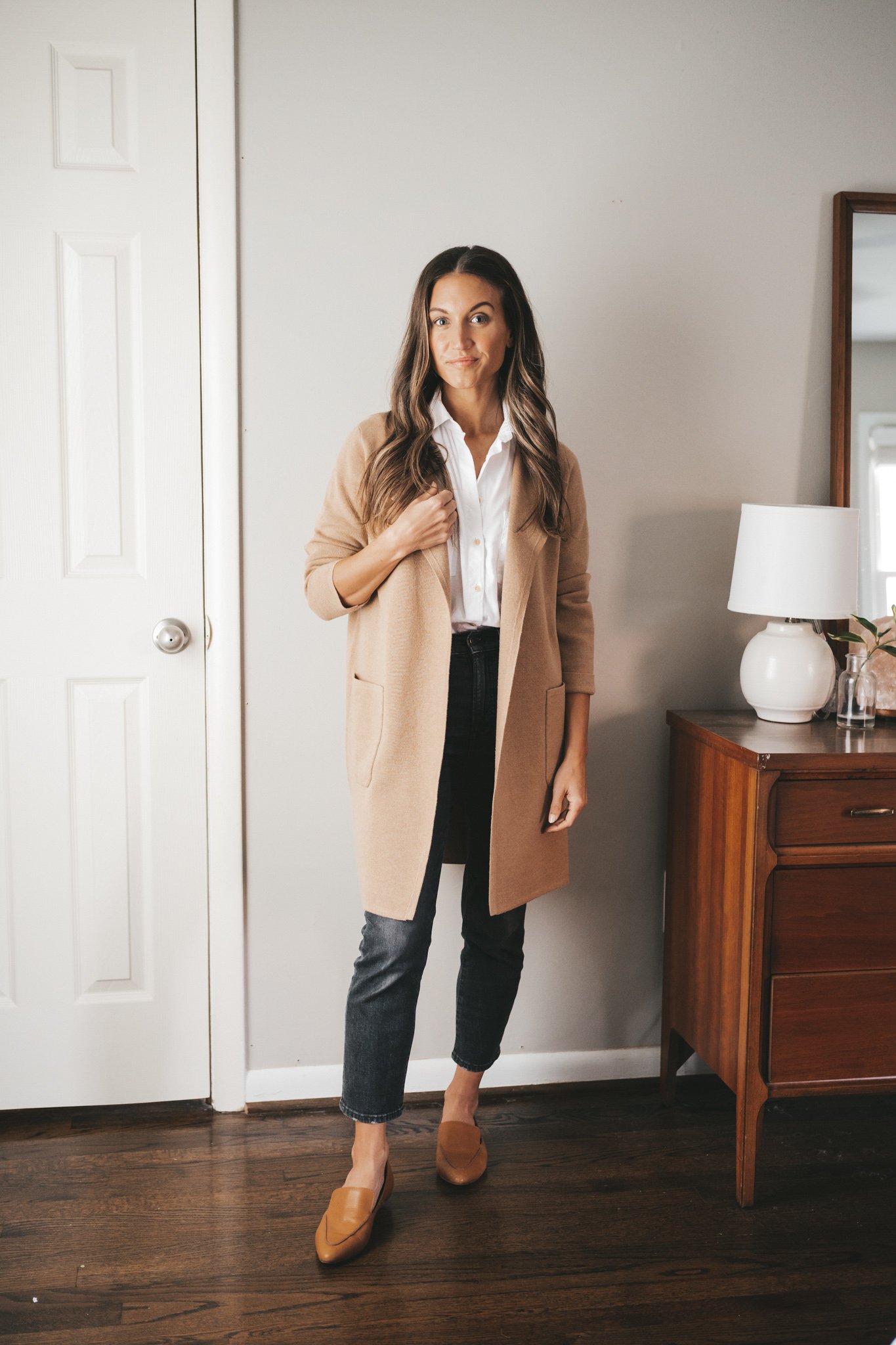 ashley wearing a white button down, tan sweater blazer, black jeans, and cognac loafers