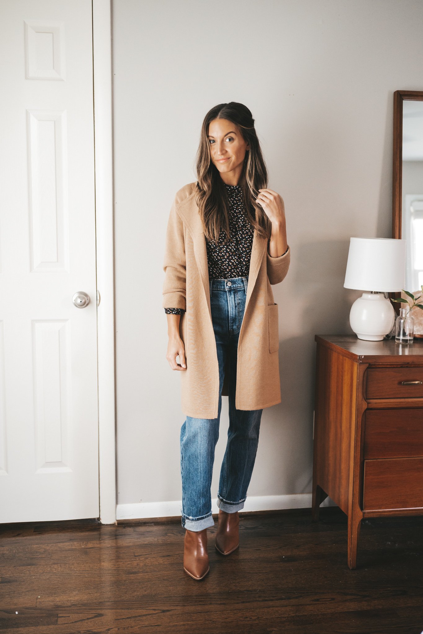ashley wearing a black floral blouse, tan sweater blazer, full length straight denim, and cognac booties